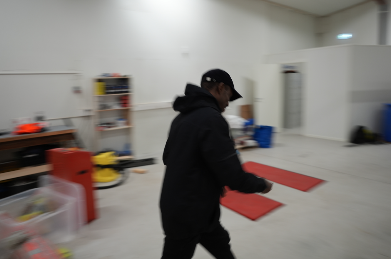 Mann i svart jakke og lue går i et rom med røde matter på gulvet og hyller med verktøy, i et industri- eller lagerlokale.