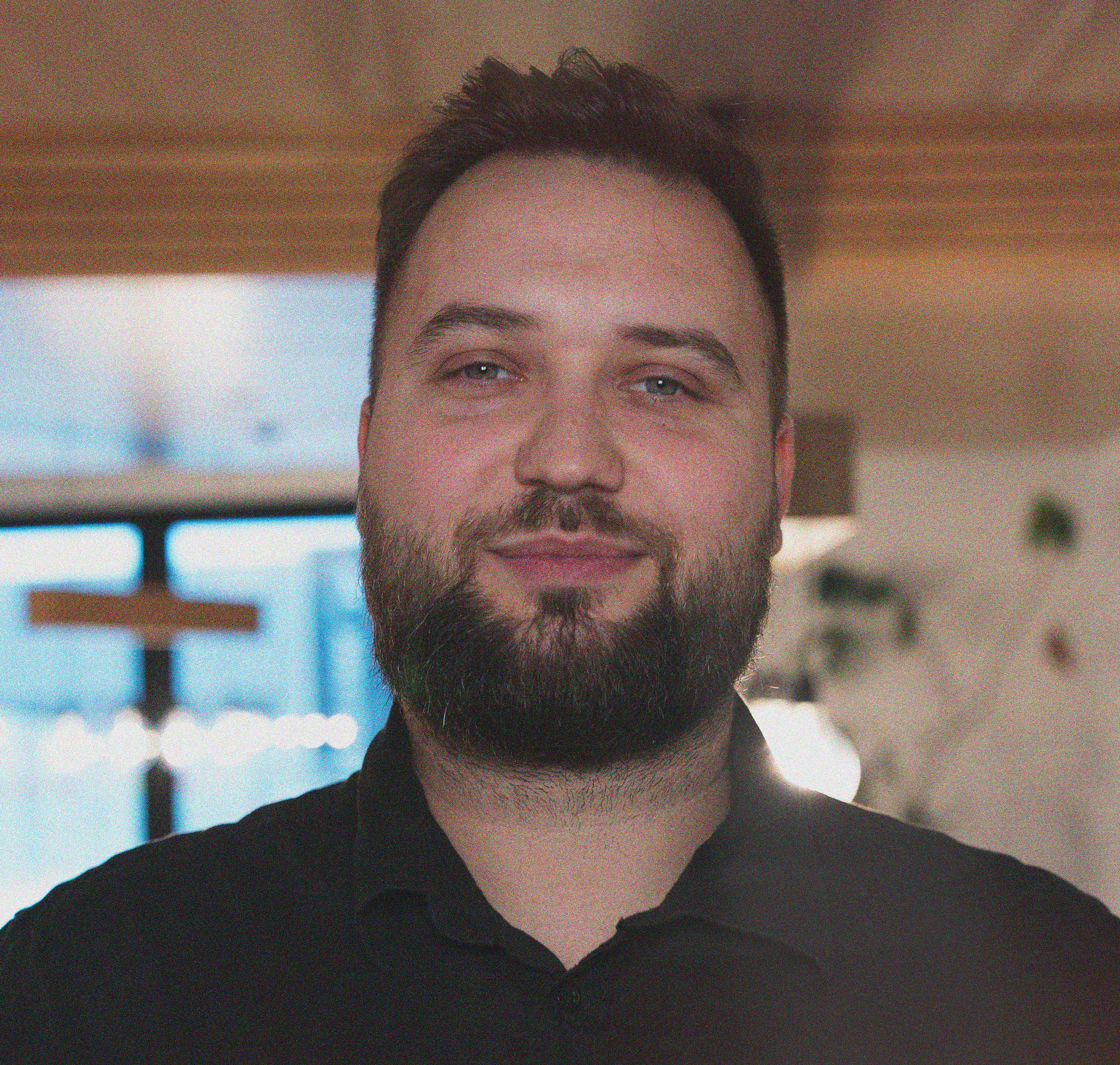 Portrett av en mann med mørk, kort hår og skjegg, iført en lys genser, stående mot en lysevegg.