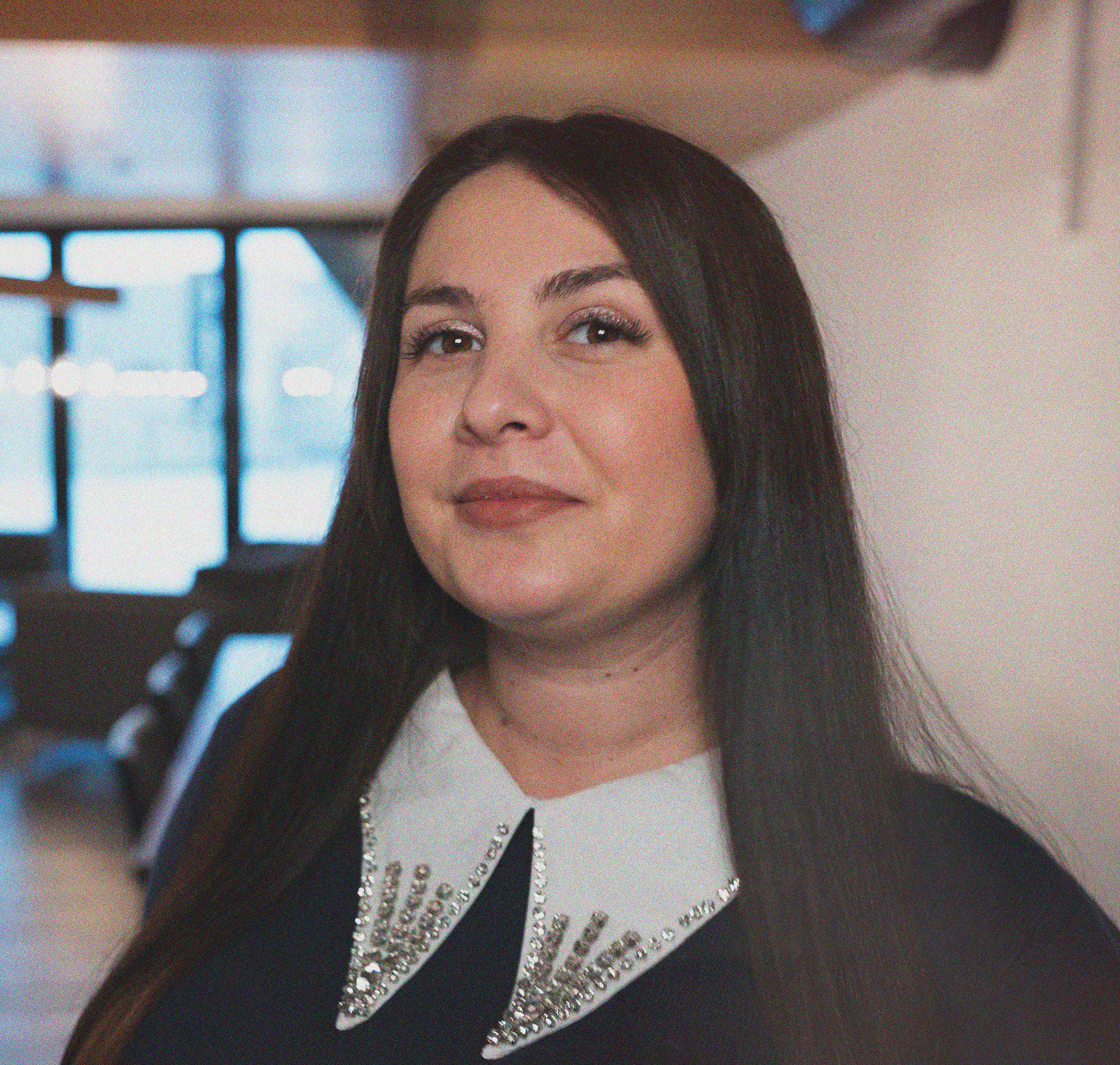 Et portrett av en kvinne med langt mørk hår, smilende med lett sminke, iført en lys genser og en delikat kjede, mot en enkel lys bakgrunn.