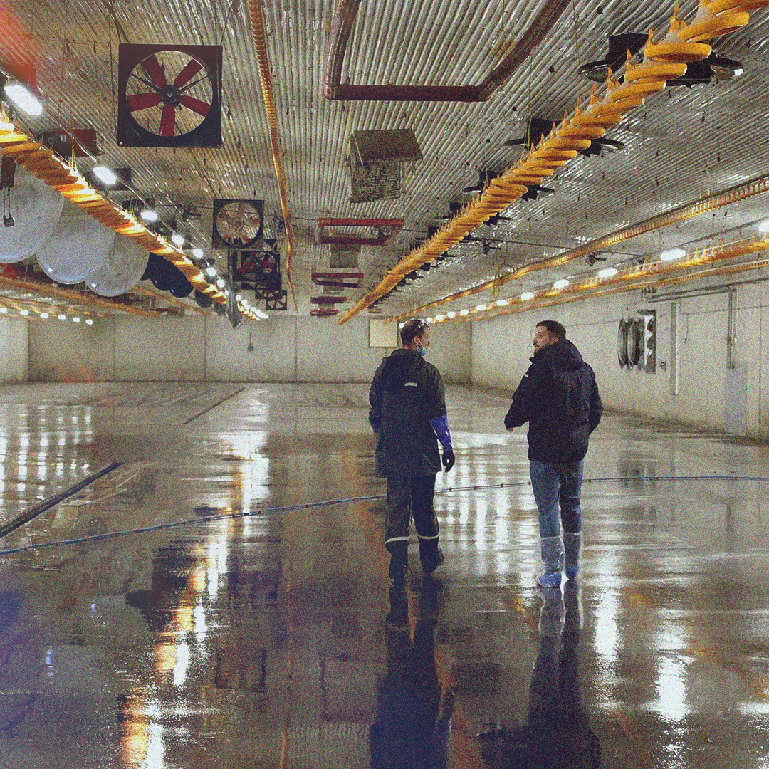 To personer i antrekk for isenkjøring eller å kommandoere, står i et stort, tomt, og fuktig industrilokale, som kan være en hangar eller en lagerhall, med våde gulv og metalltak.