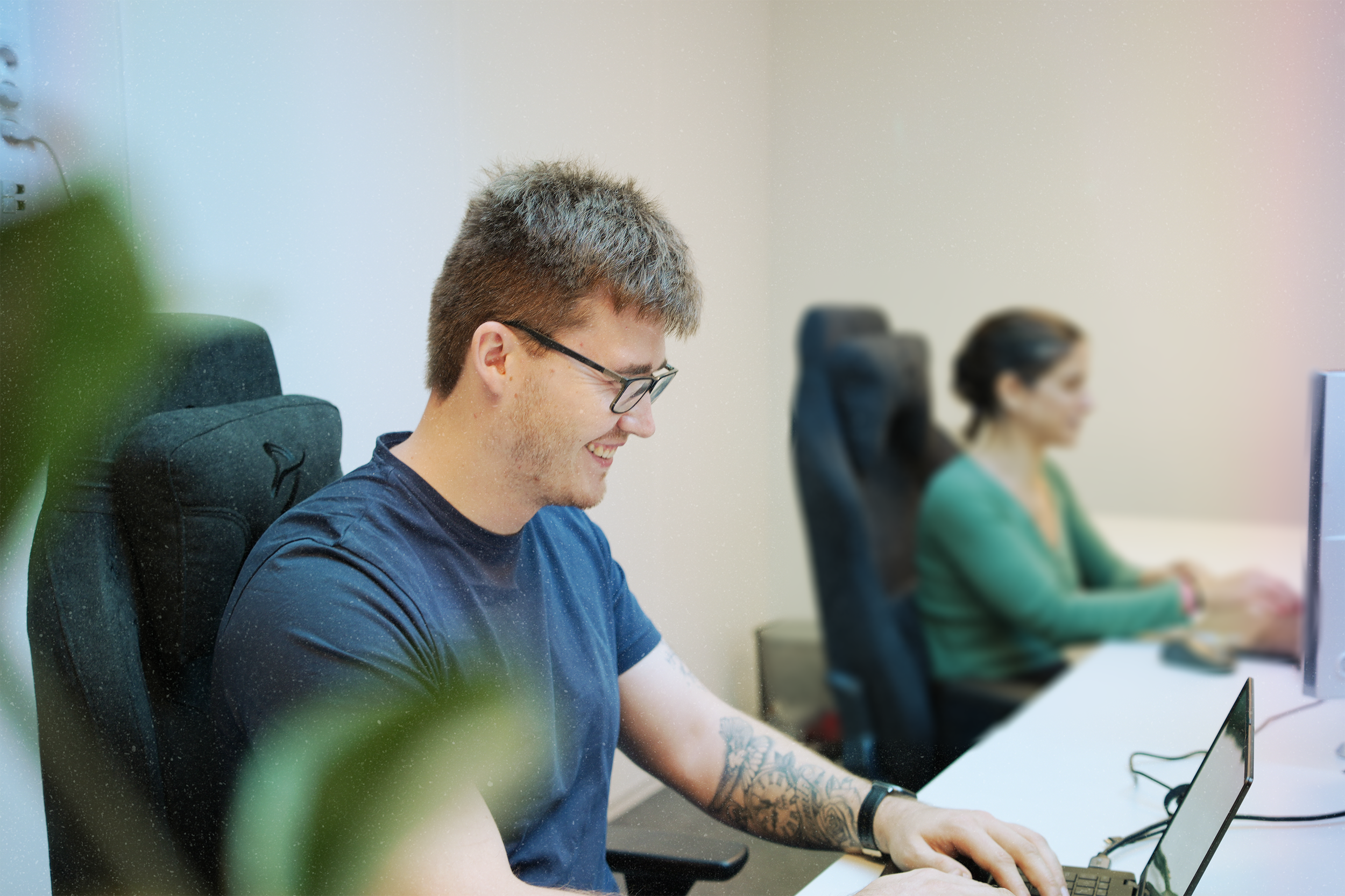 To personer jobber på hver sin datamaskin på kontor, en mann med briller og tatovering smiler mens han bruker en bærbar datamaskin, en kvinne sitter i bakgrunnen.