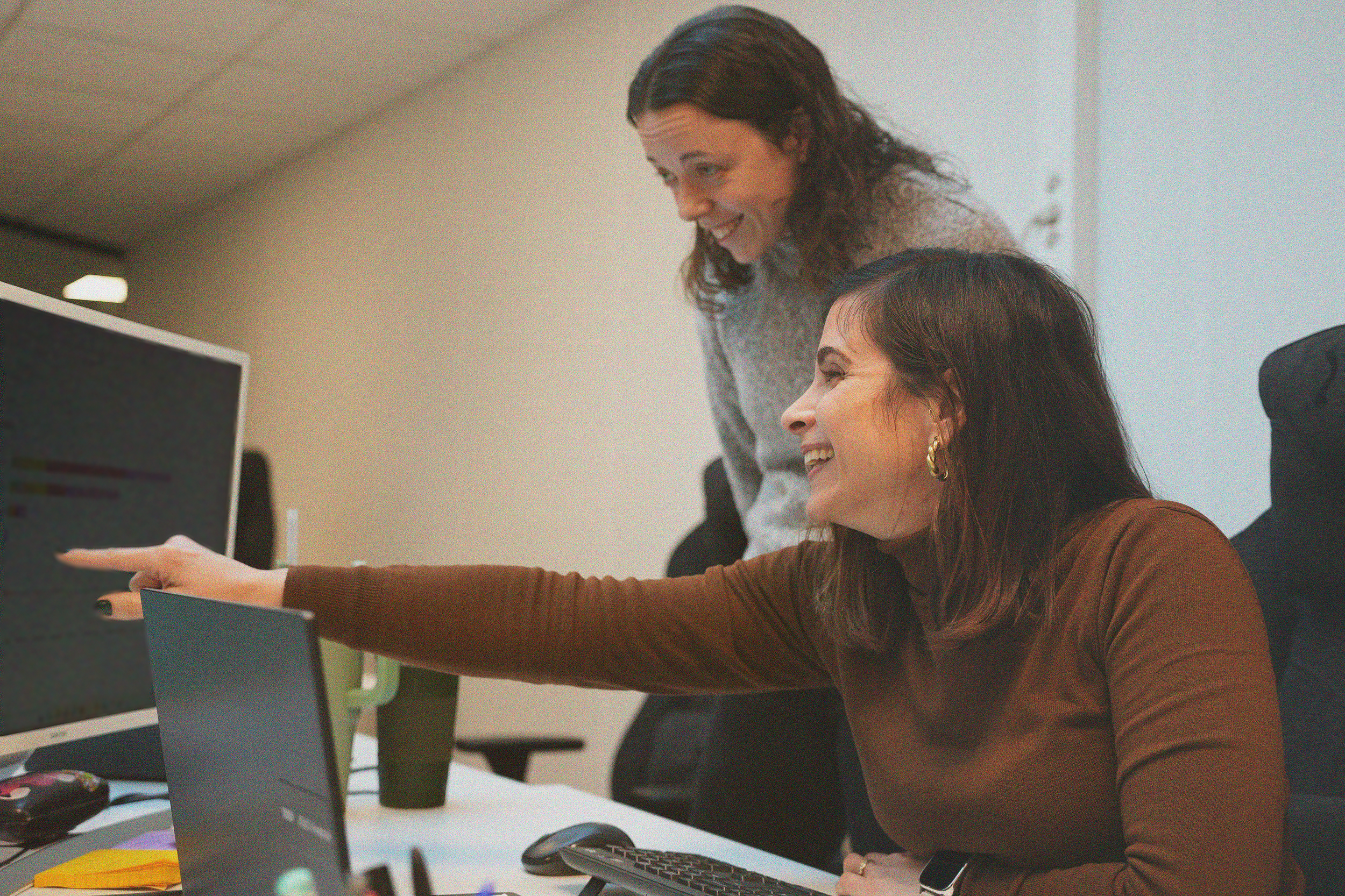 To kvinner i et kontor, den ene peker på en skjerm, de smiler og ser engasjerte ut.