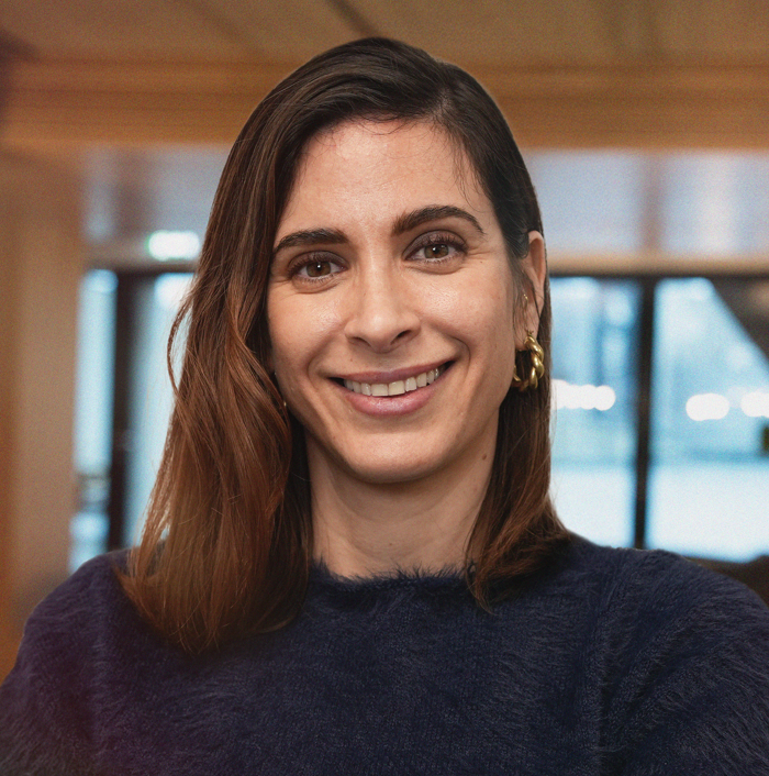 Et bilde av en smilende kvinne med brune øyne, mørkt hår trukket tilbake, og iført en lilla strikket genser, med grønne planter i bakgrunnen.