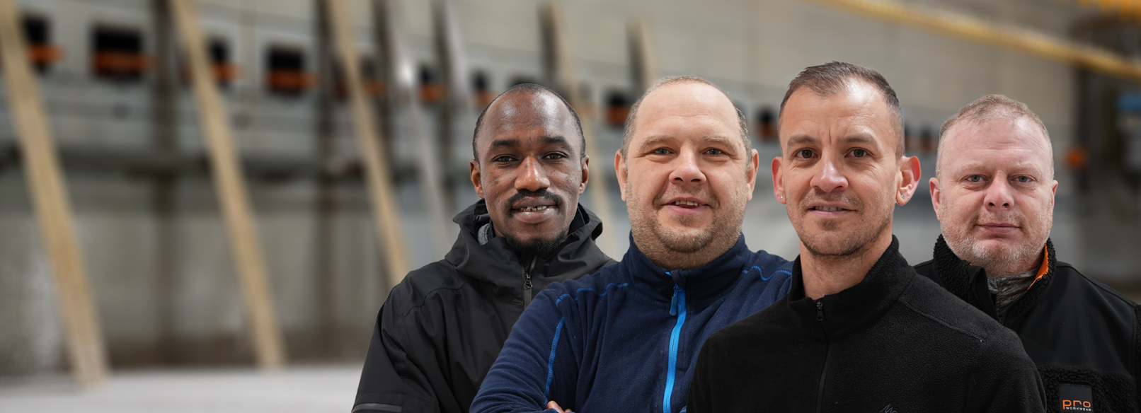 Fire menn står inntil hverandre i en lagerbygning, alle smilende, kledd i trenings- eller utendørsarbeidstøy.