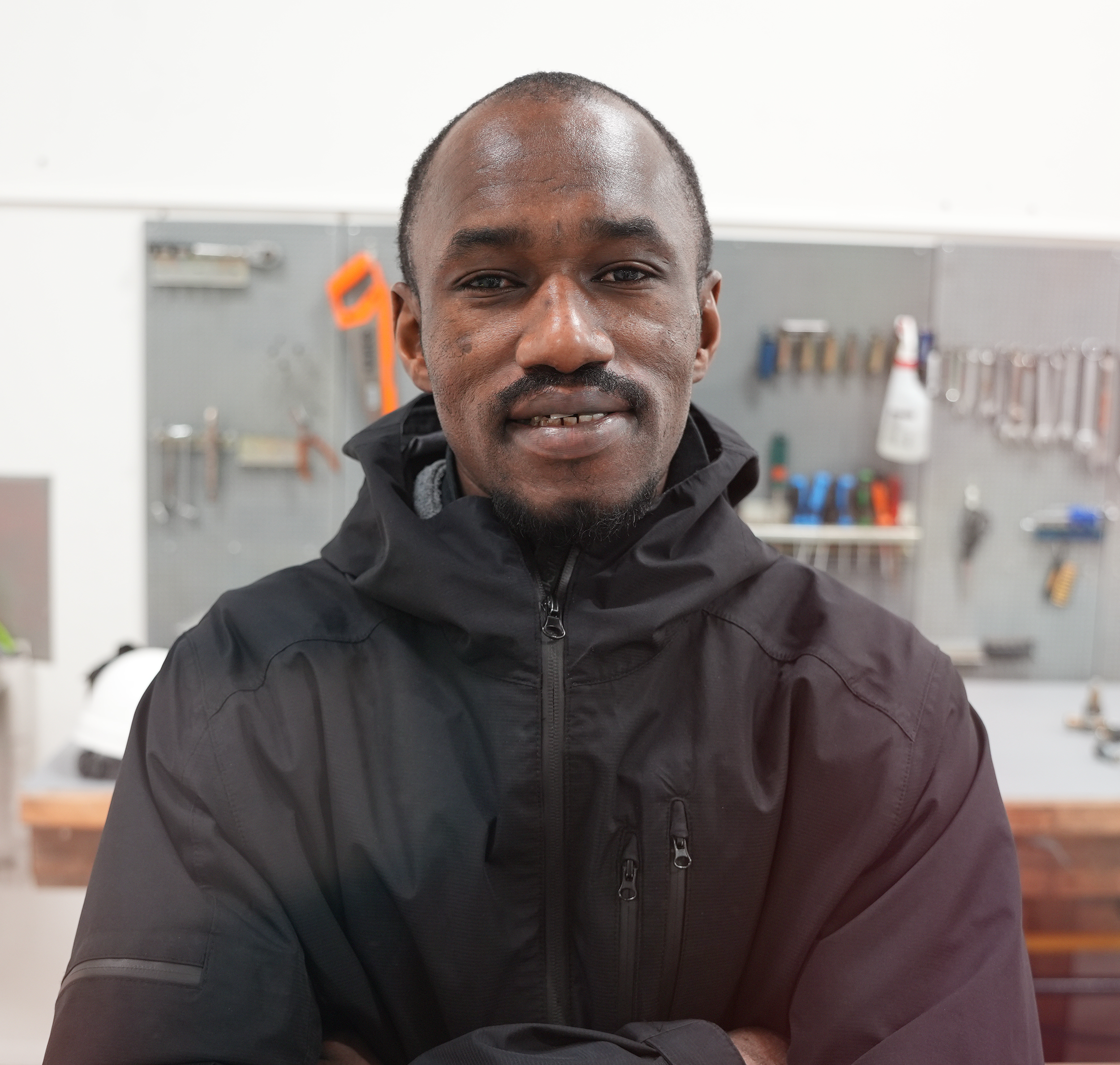 En mann med mørk hud og kort håret, iført svart jakke, står i en verksted med verktøy på veggen i bakgrunnen.