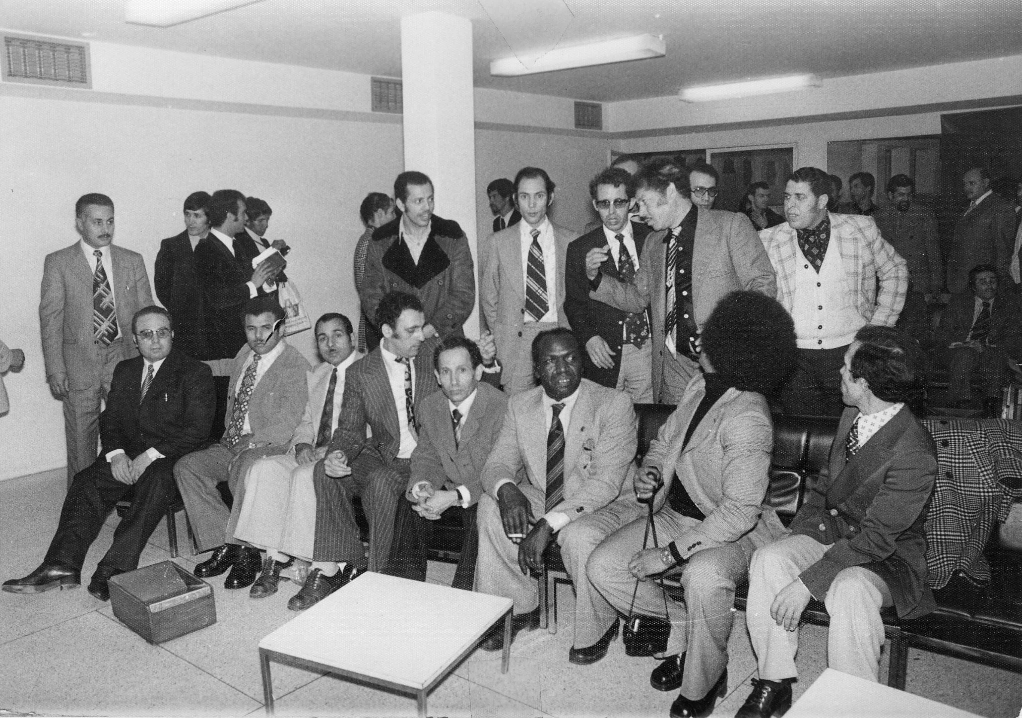 Gruppe mennesker i forretningsantrekk samles i et rom, noen sitter og noen står, i svart-hvitt fotografi.