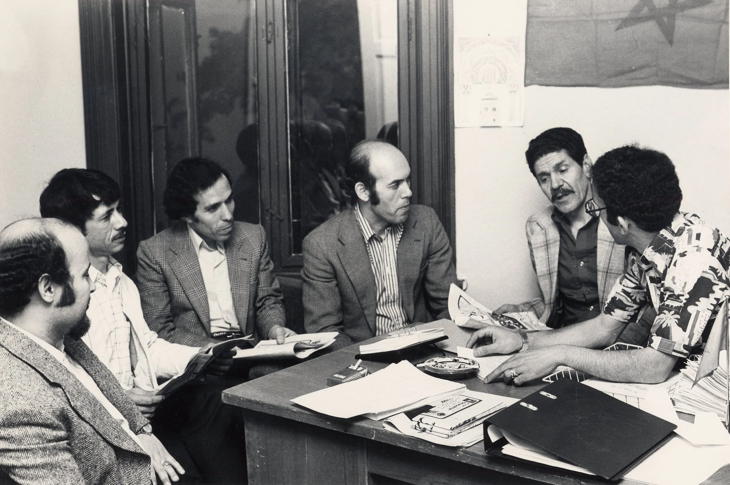 En gruppe menn er samlet rundt et bord i et møte, de diskuterer og har aviser og dokumenter foran seg.