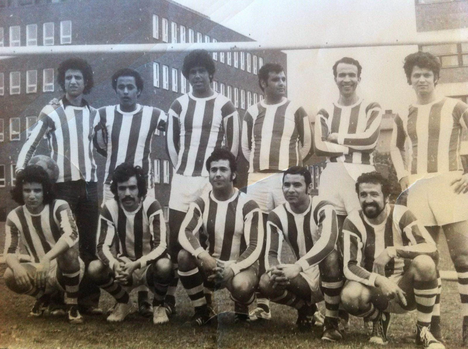 En svart-hvitt gruppefotografi av fotballspillere i stripedoseringsdrakter, tatt utenfor en bygning.