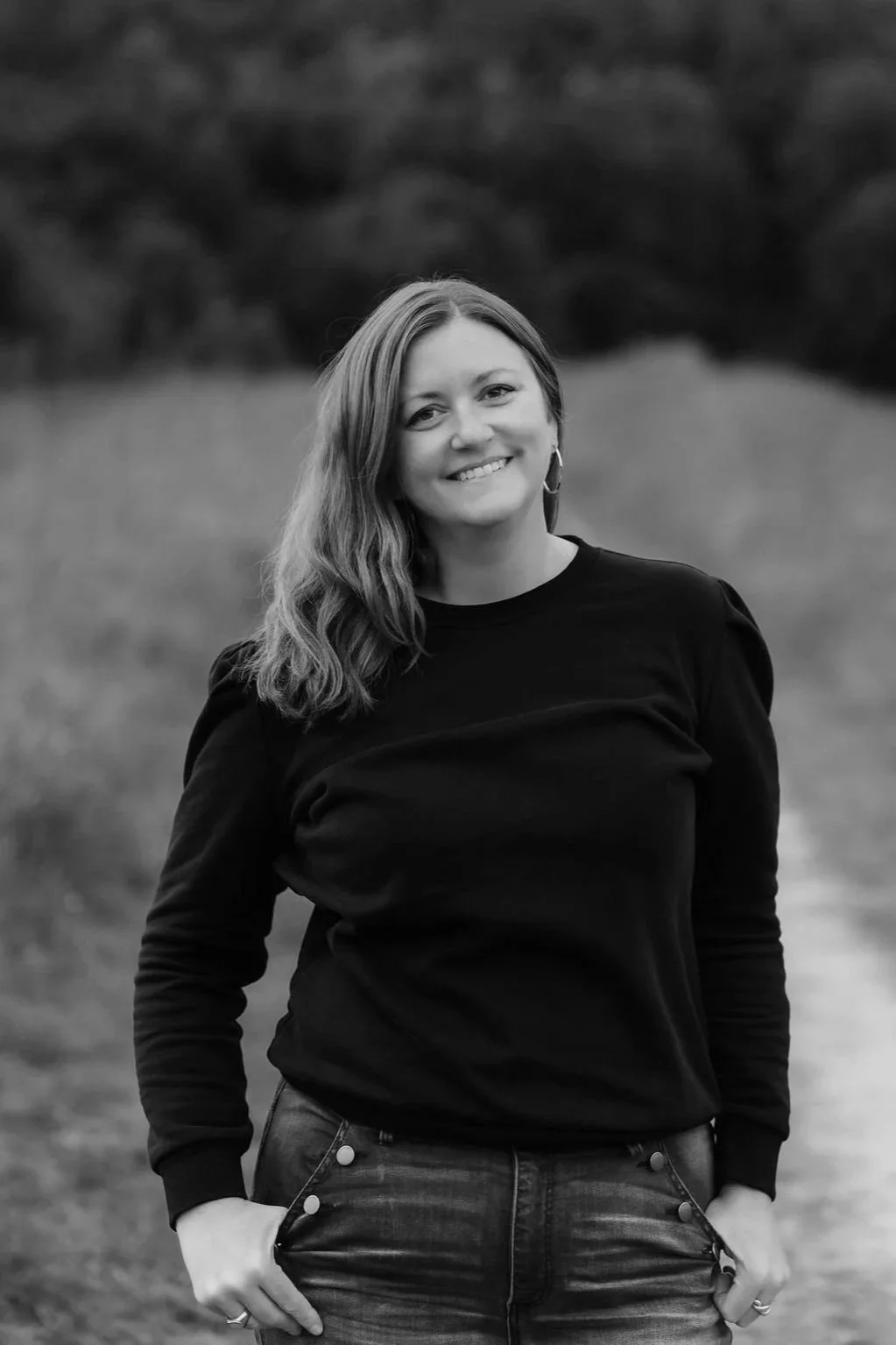 A woman with shoulder-length hair, wearing a long-sleeved black shirt and jeans, smiling outdoors in a natural setting.
