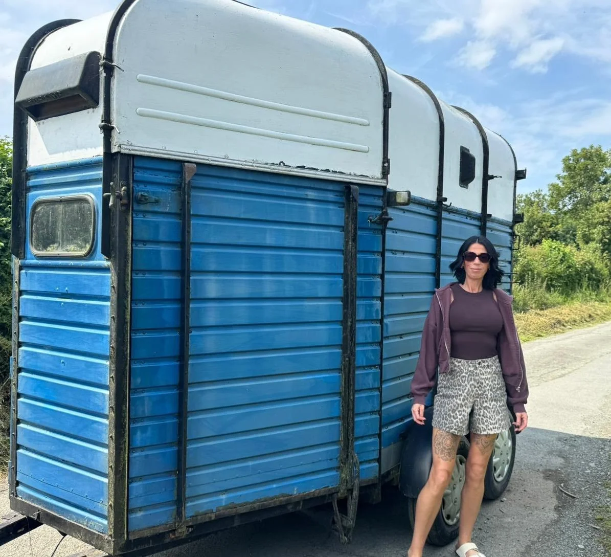 A woman wearing sunglasses, a purple jacket, a black top, and leopard print shorts standing beside a converted blue and white trailer on a rural road with greenery and a partly cloudy sky in the background.