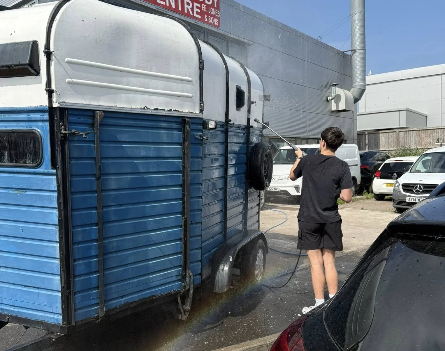 A person washing a blue and white food truck in a parking lot with several parked cars and industrial buildings in the background.