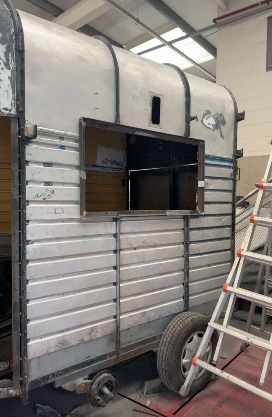 Metal trailer with an opening window and a horse silhouette, on wheels, in a warehouse or workshop setting with a ladder nearby.
