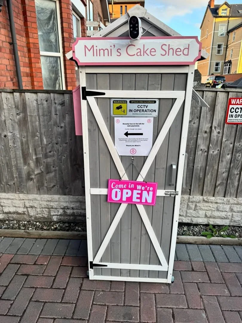 A small grey wooden shed named 'Mimi's Cake Shed' with a pink and white sign that says 'Come in We're OPEN.' The shed has a CCTV camera mounted above the sign and is in front of a wooden fence, with a brick building and colorful houses in the background.