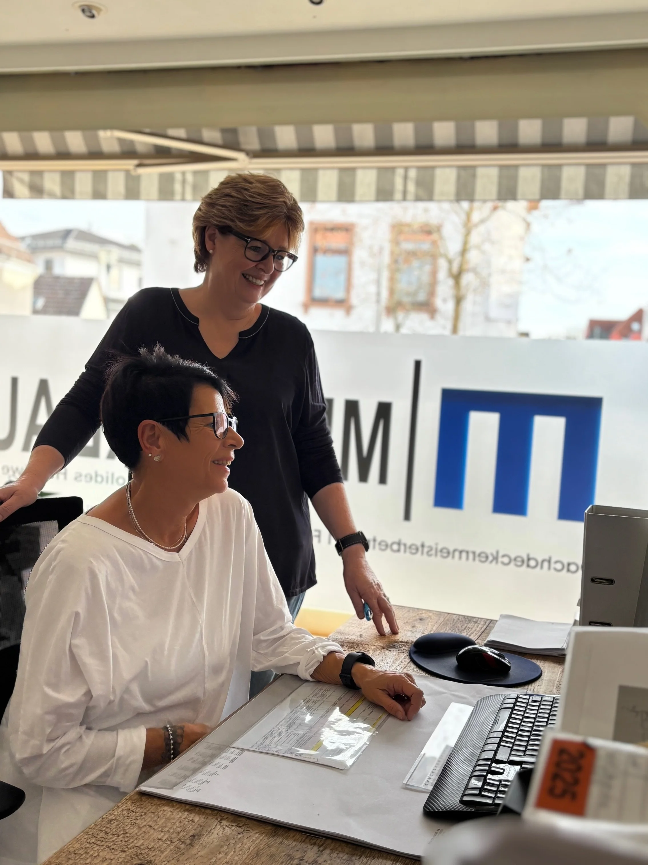 Zwei Frauen, eine sitzend, die andere stehend, lächeln in einem Büro vor einem Computer.