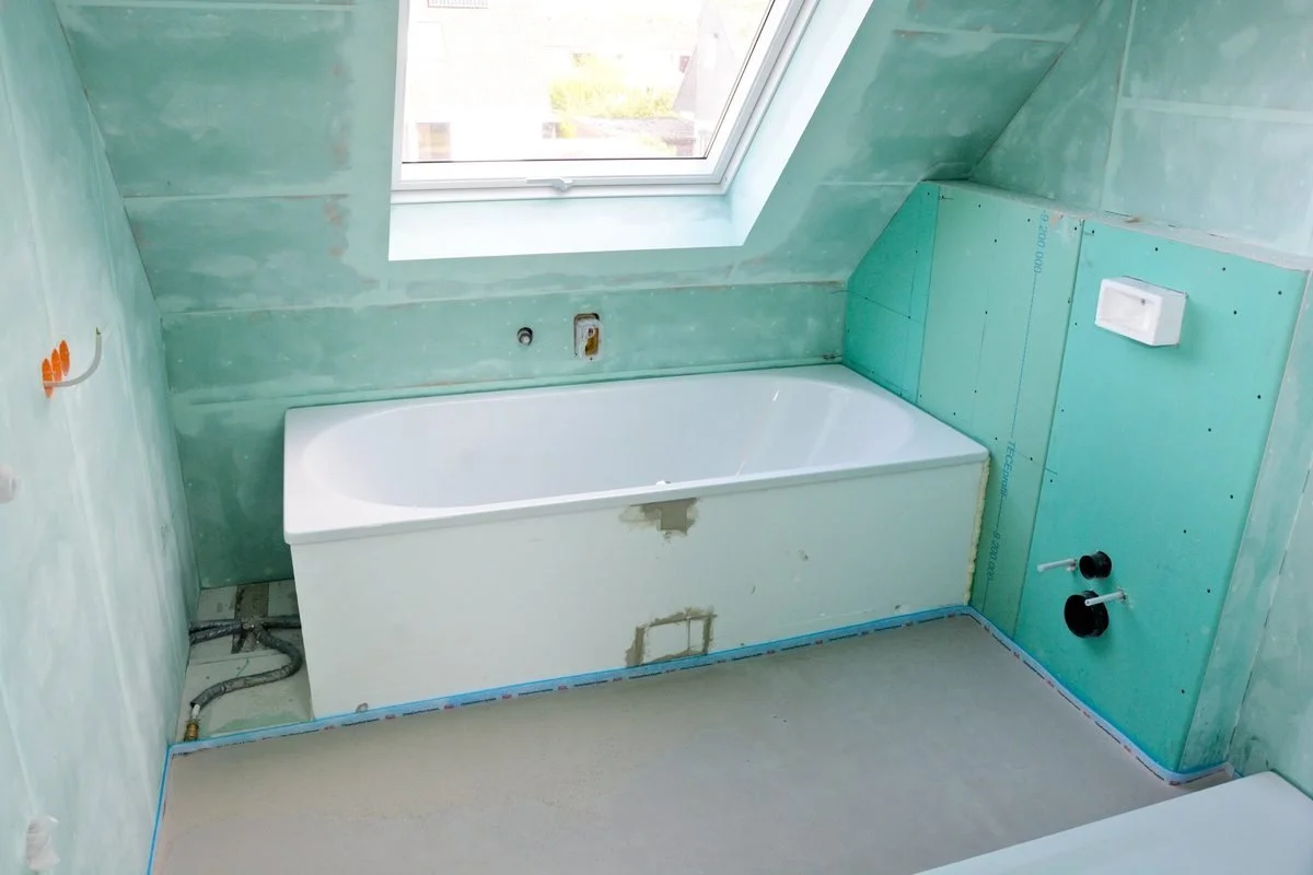 Badezimmer im Bau, mit Badewanne, unbehandelten Wänden und Rohren, Fenster an der Decke, und Installationsausschnitten.