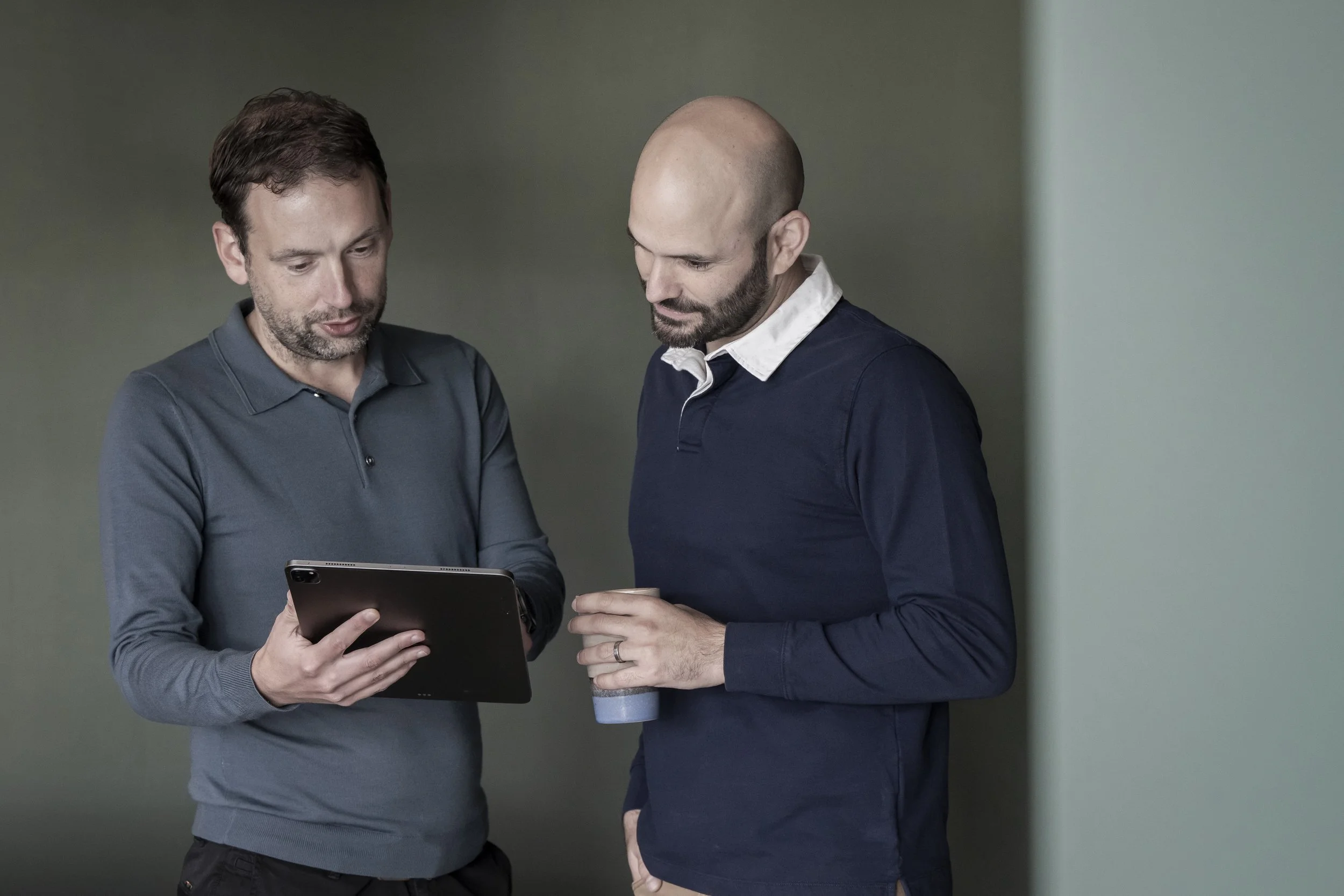 Two men stand side by side, looking at a tablet device together, one holding a cup of coffee.