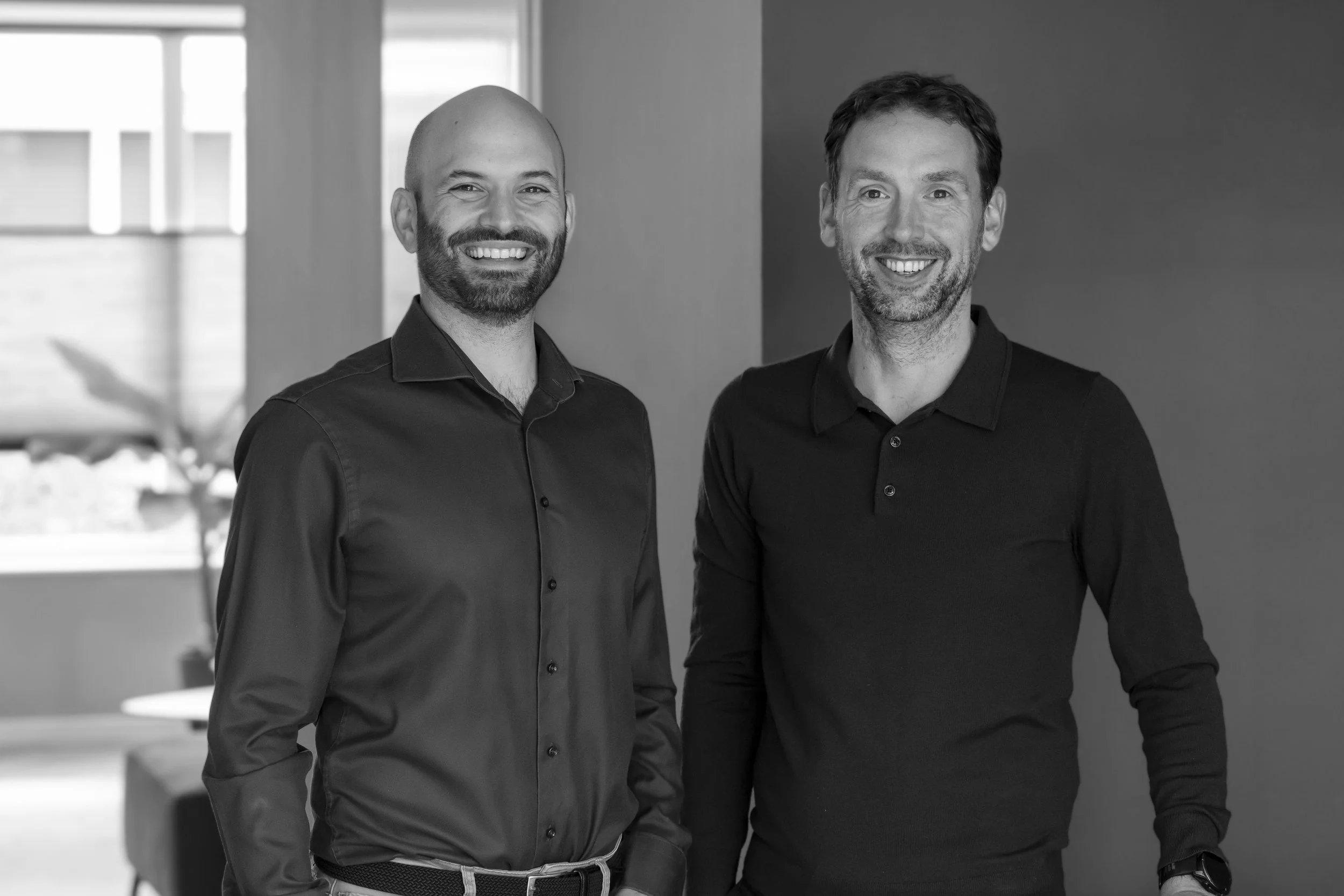 Two smiling men standing side by side indoors with a window and a plant in the background.