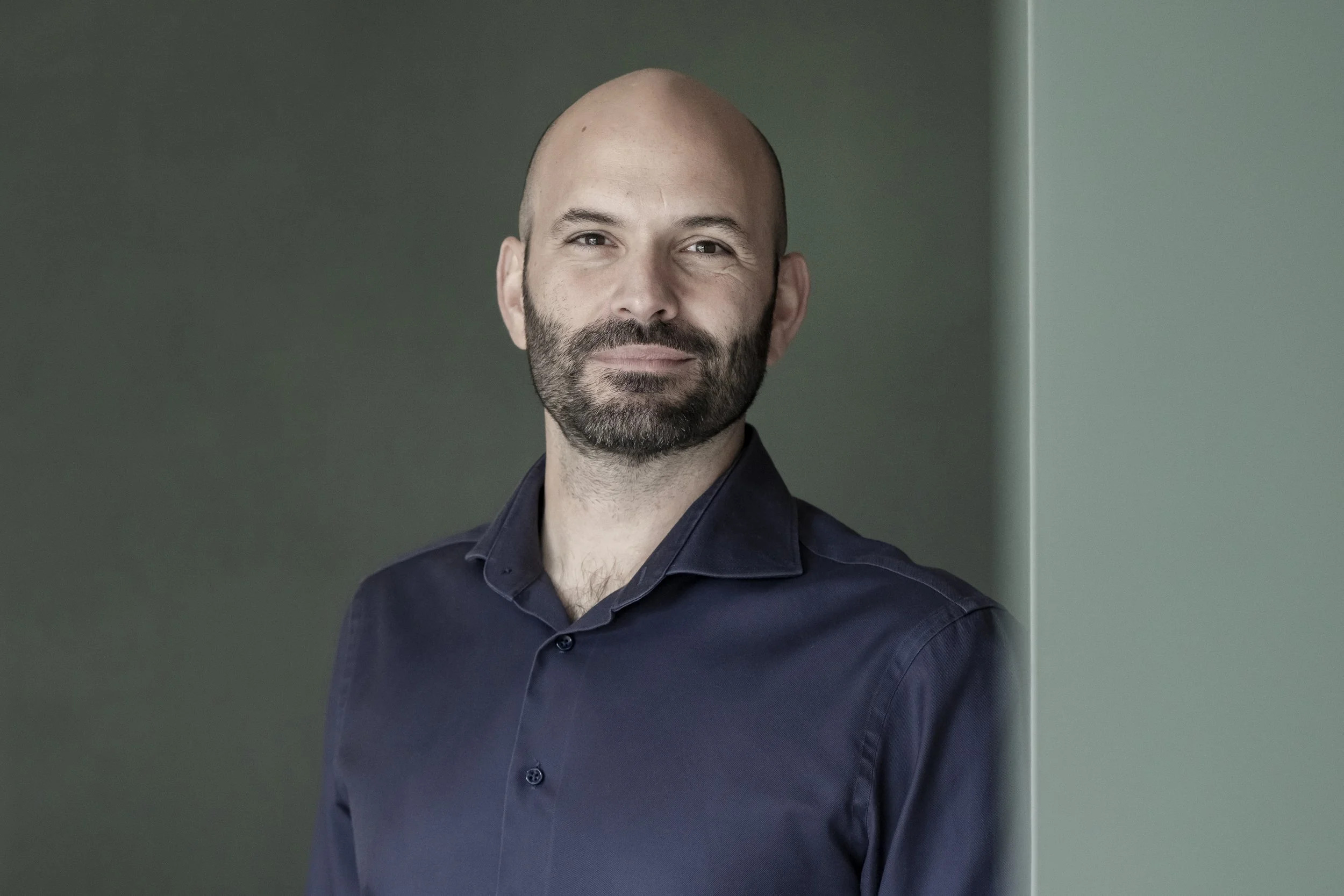 A man with a beard and bald head, wearing a dark button-up shirt, standing against a plain color background, looking confidently into the camera.