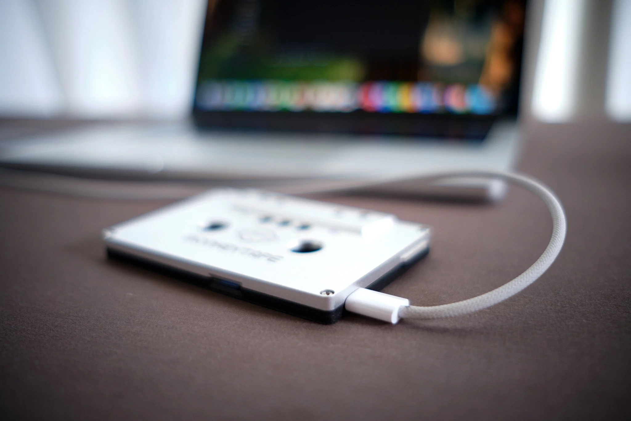 A vintage black cassette tape plugged into a modern silver laptop on a wooden desk