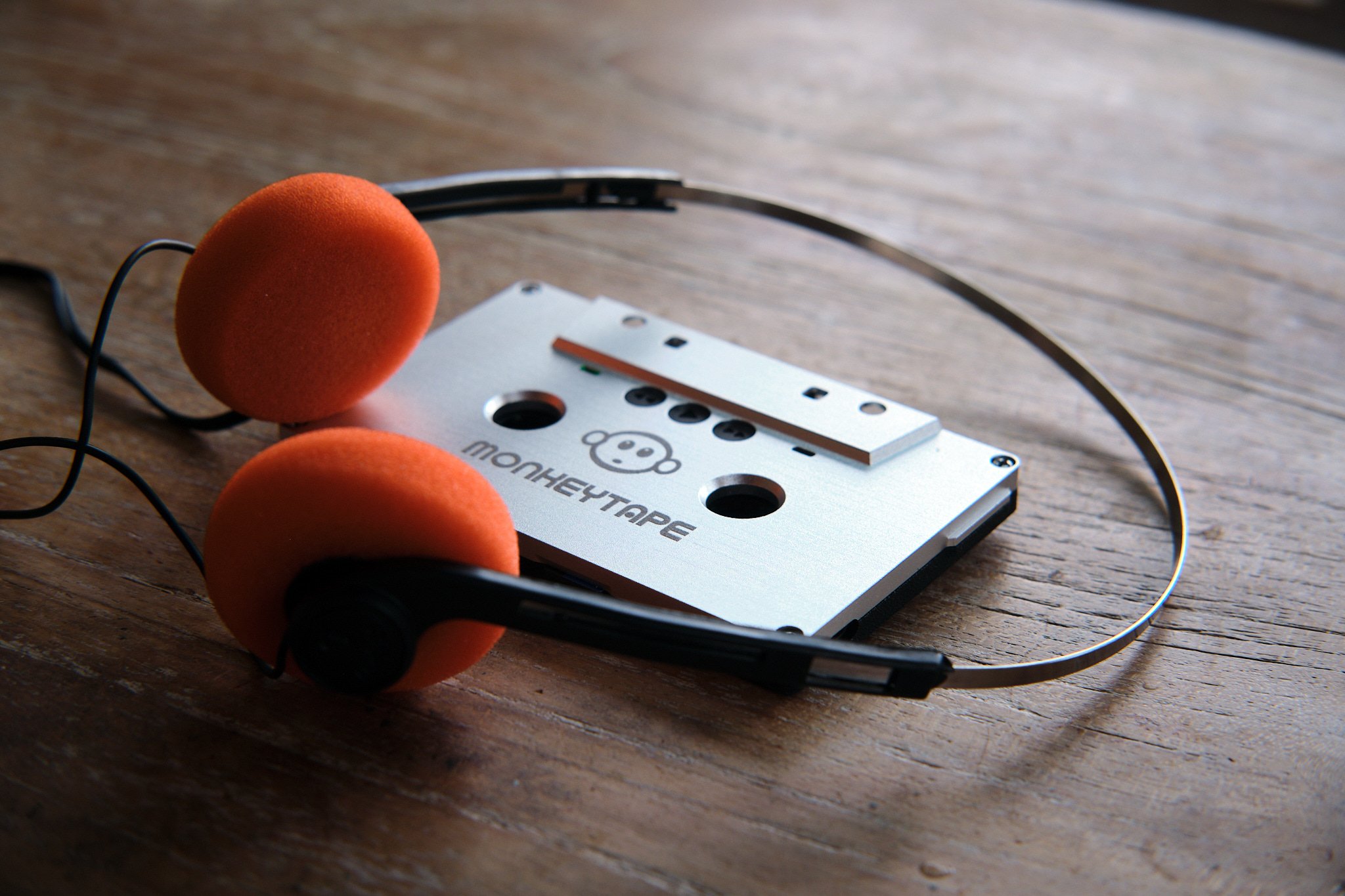 A silver cassette tape labeled 'MonkeyTape' with a monkey face logo, placed on a wooden surface with headphones in the background.