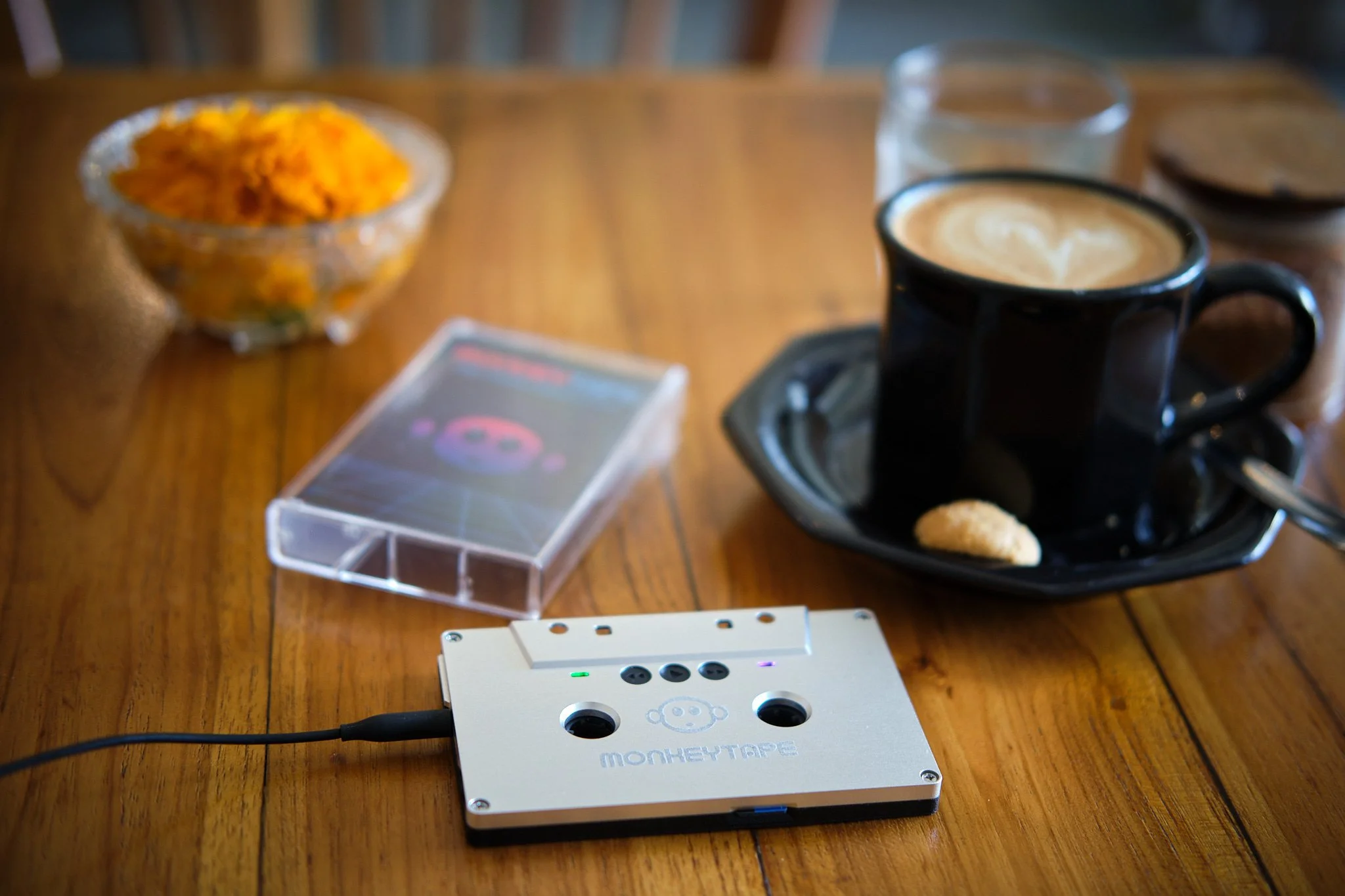 A wooden table with a black coffee cup on a saucer, a small snack, a glass of water, a bowl of food, a portable electronic device, and a small electronic gadget labeled 'MonkeyTape'.