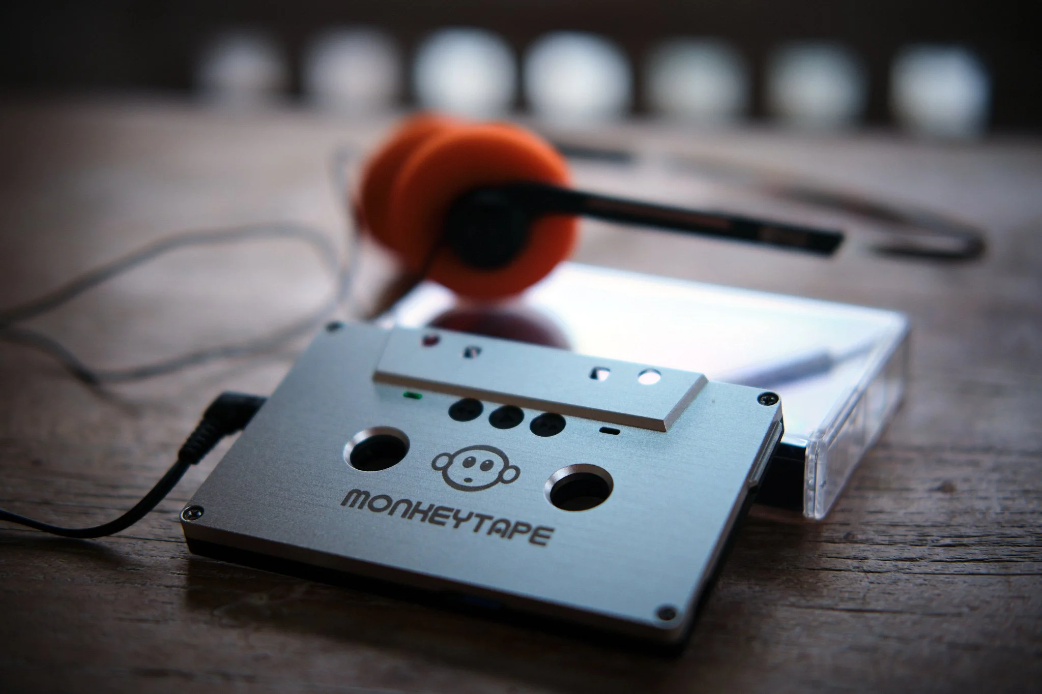 A vintage metallic cassette tape with a monkey face logo and the word 'MonkeyTape' on it, placed on a wooden surface with an orange headphone and a clear plastic case in the background.