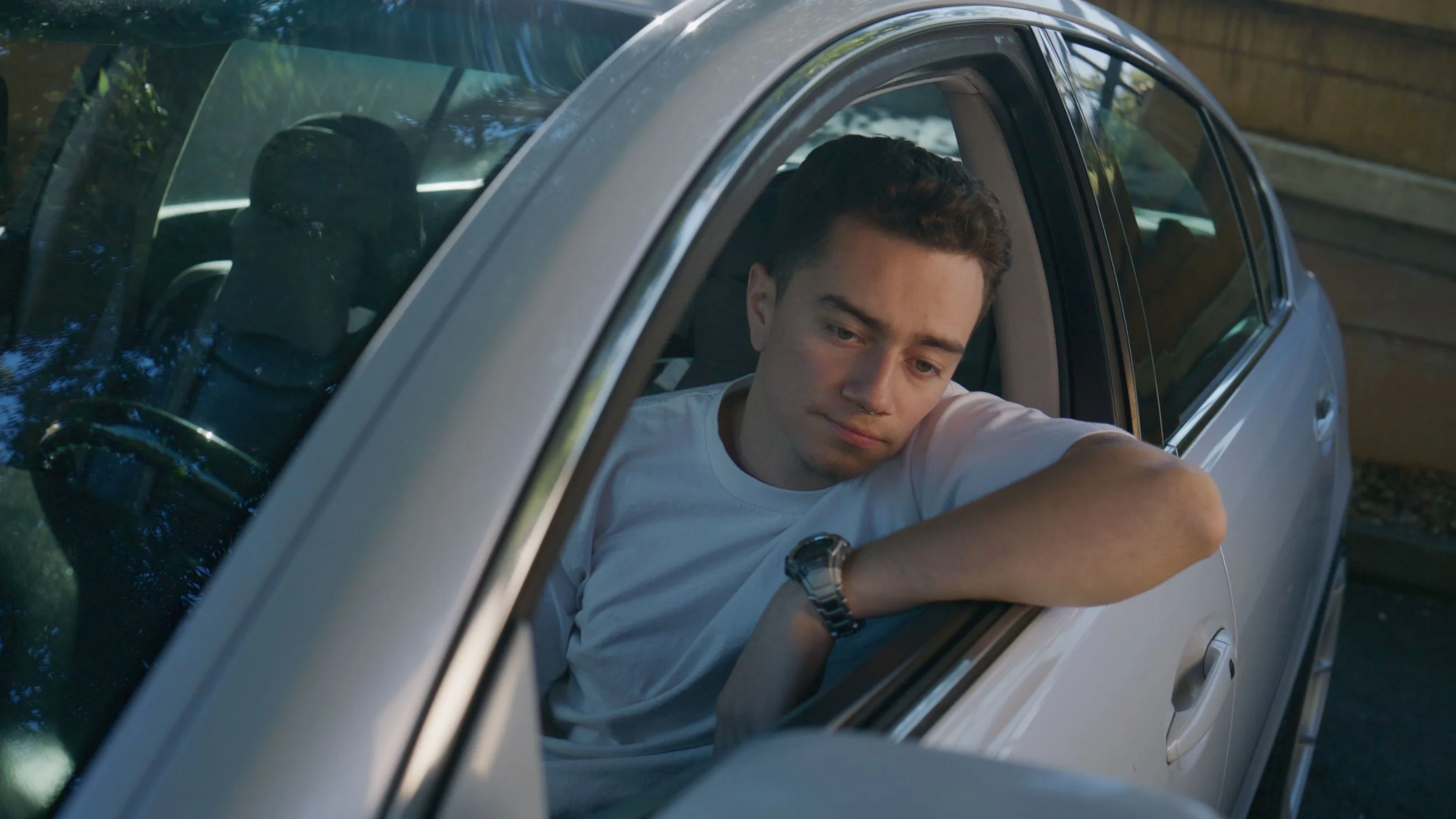 A young man with short dark hair looks out of his car window with a sad or contemplative expression, resting his arm on the window frame. The car is parked outside near a wooden fence.