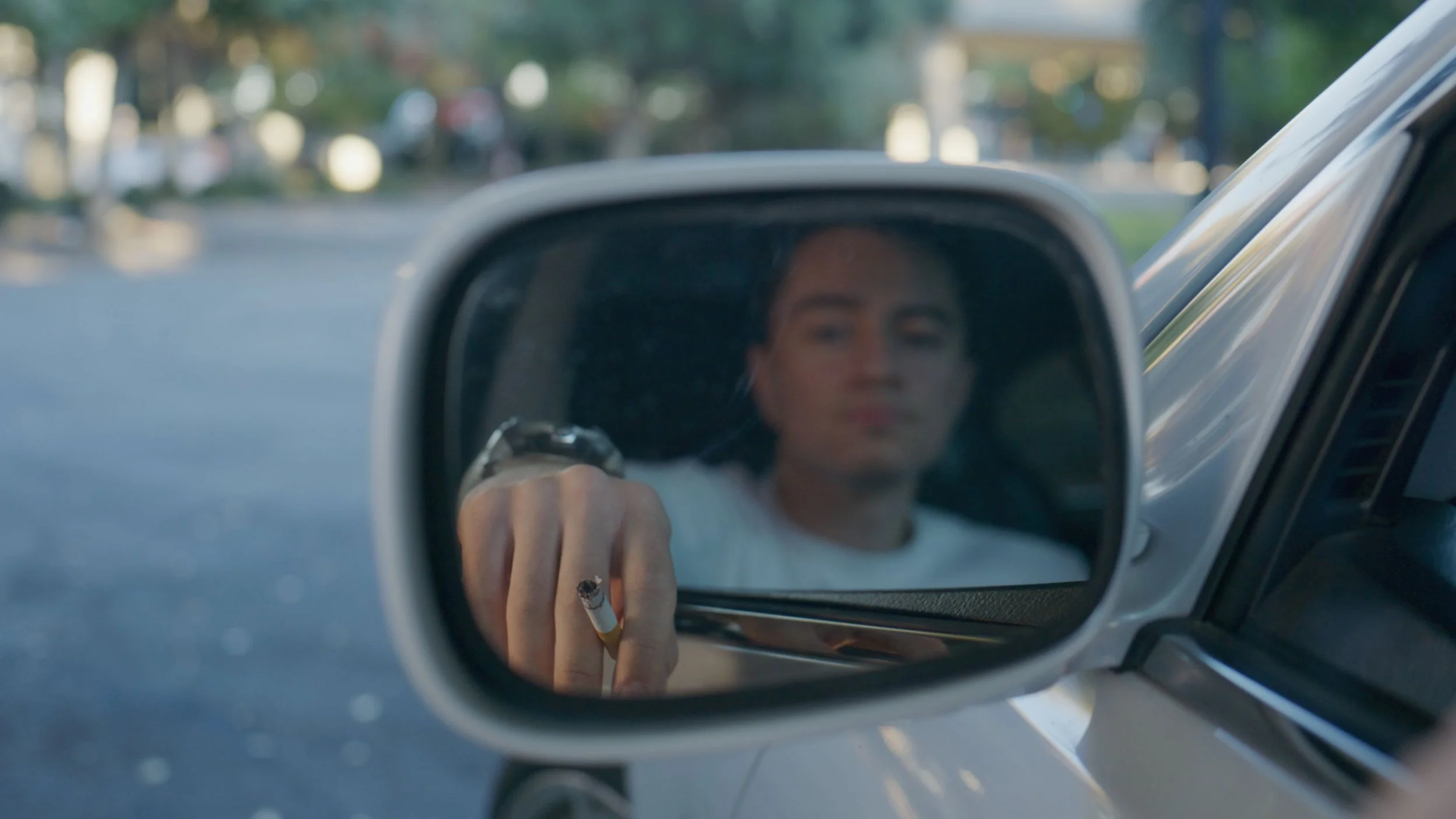 A person is seen in the reflection of a car's side mirror, holding a cigarette.