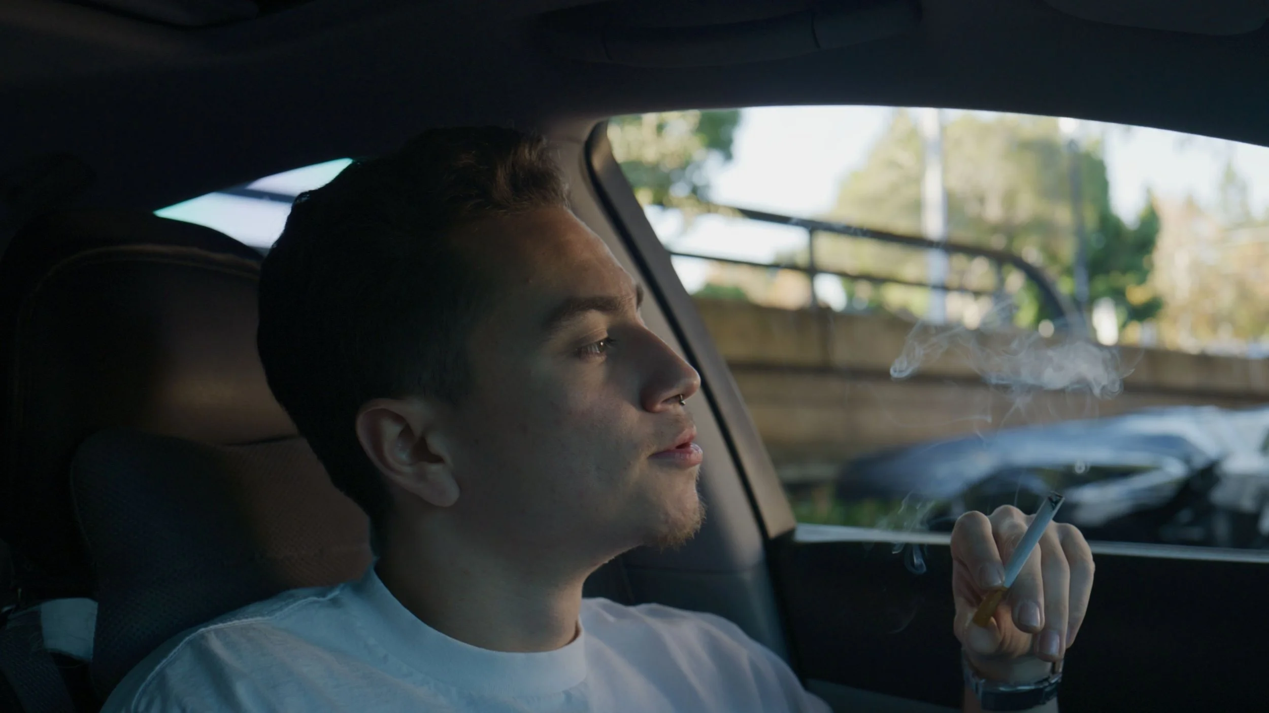 A young man with dark hair and a nose ring, sitting in a car, smoking a cigarette with smoke rising, with daylight outside visible through the window.