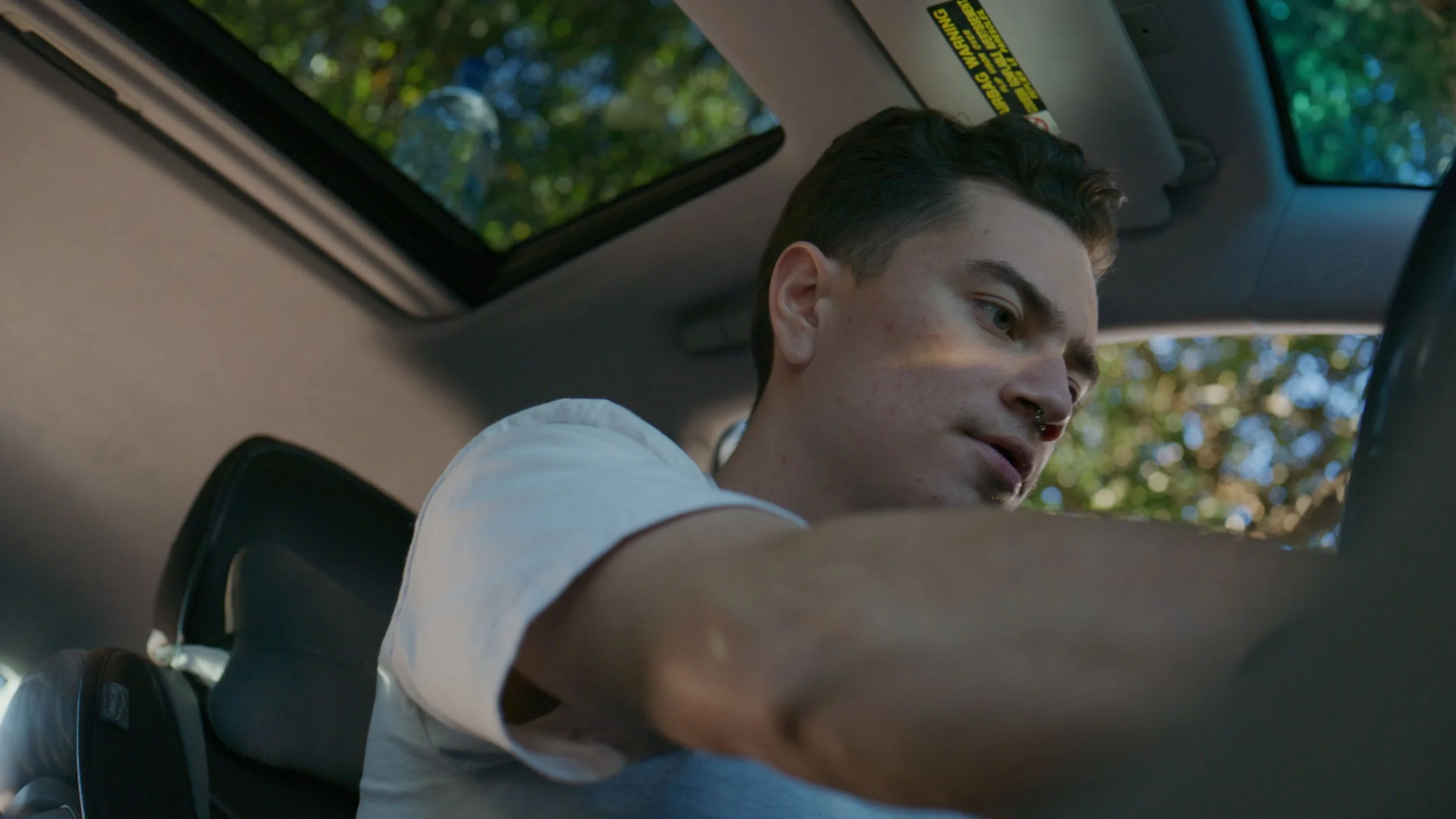 A young man with dark hair, wearing a white t-shirt, is sitting inside a car and reaching through the open window, with trees and blue sky visible outside.