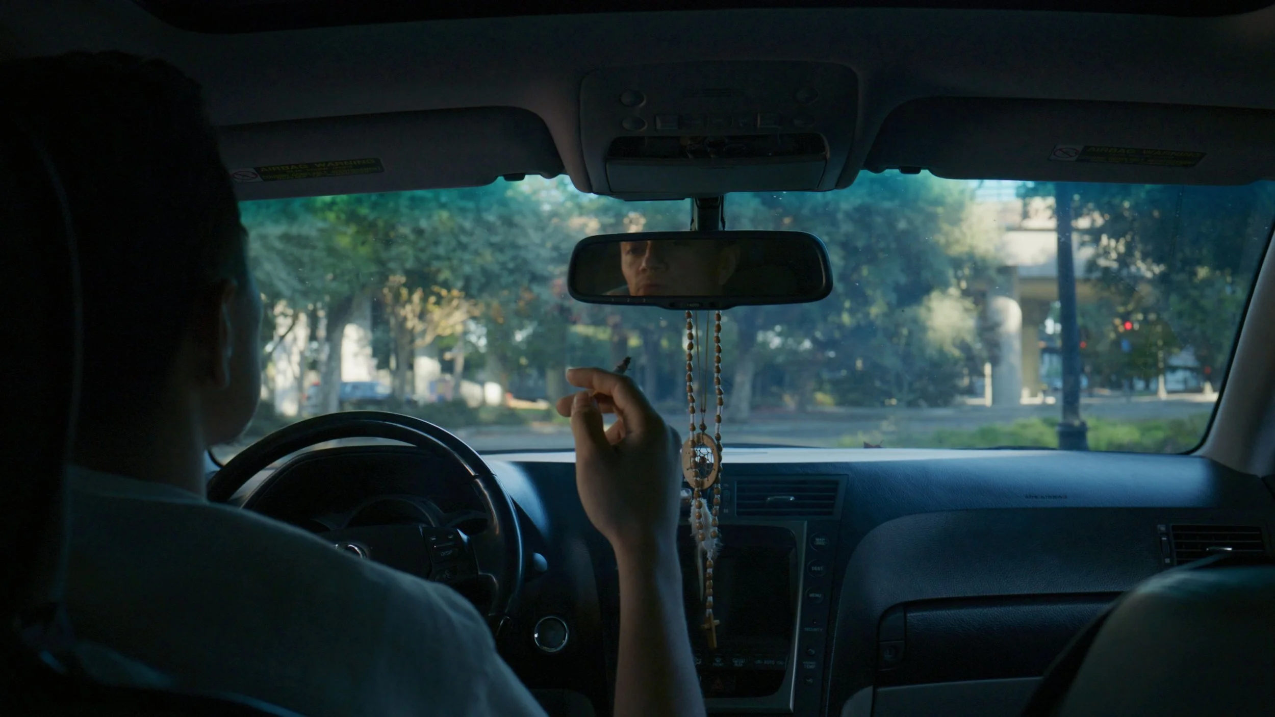 Person sitting in the driver's seat of a car, holding a cigarette, with a rearview mirror hanging with prayer beads and a spiritual pendant, outside view of trees and buildings through the windshield.