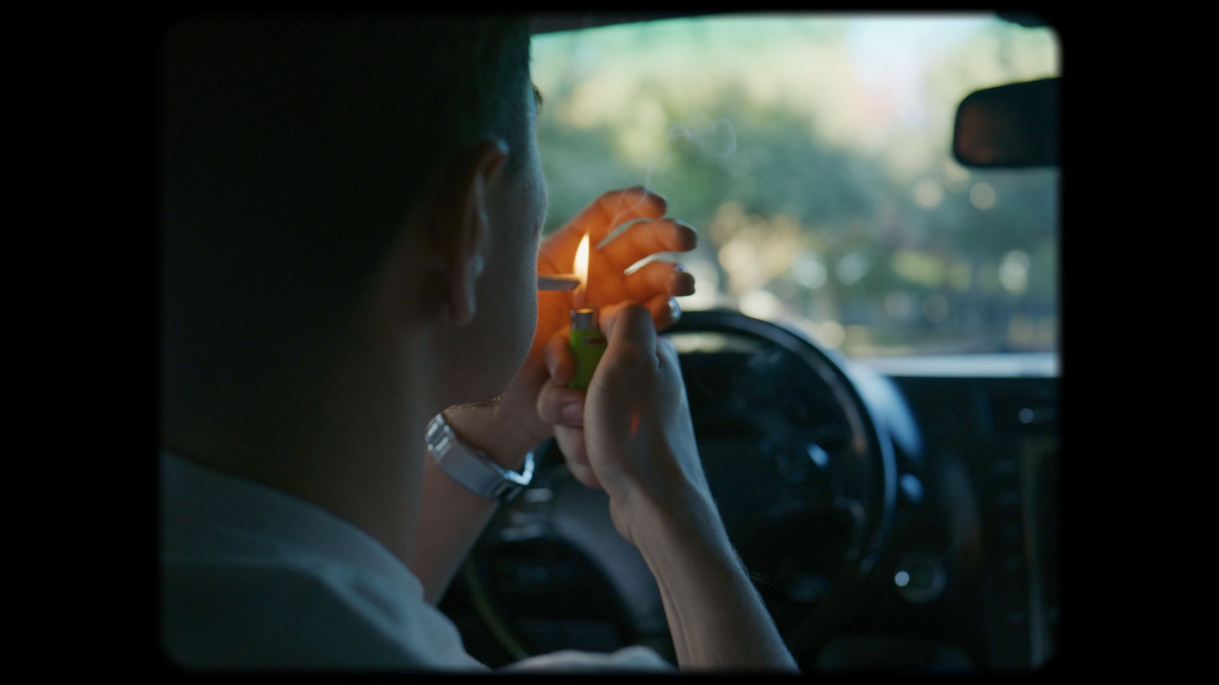 A person in a car, lighting a cigarette with a lighter, with a view of the steering wheel and dashboard visible.