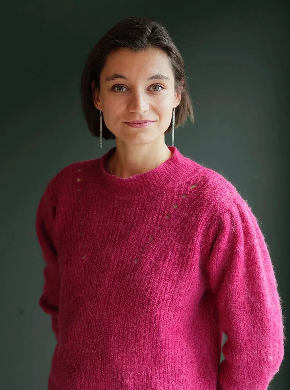 A woman with short dark hair wearing a pink sweater with small holes and long earrings, standing against a dark green background.