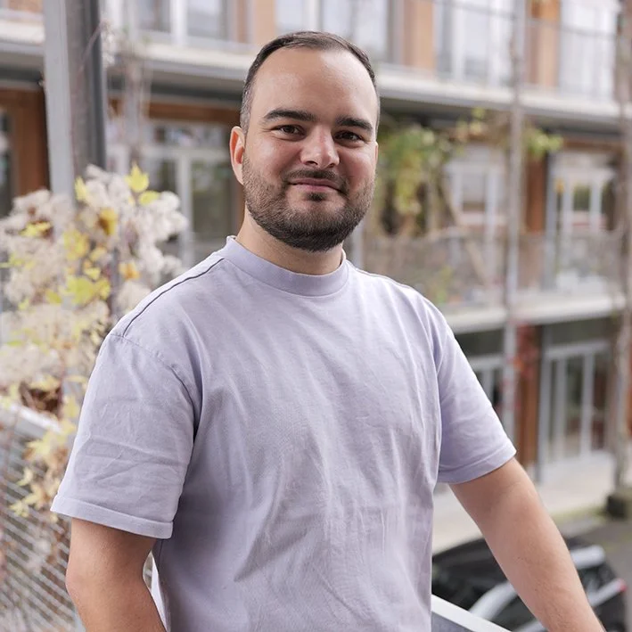 A man with a beard and short hair stands outdoors on a balcony or terrace, wearing a light gray T-shirt, with a building and some plants in the background.