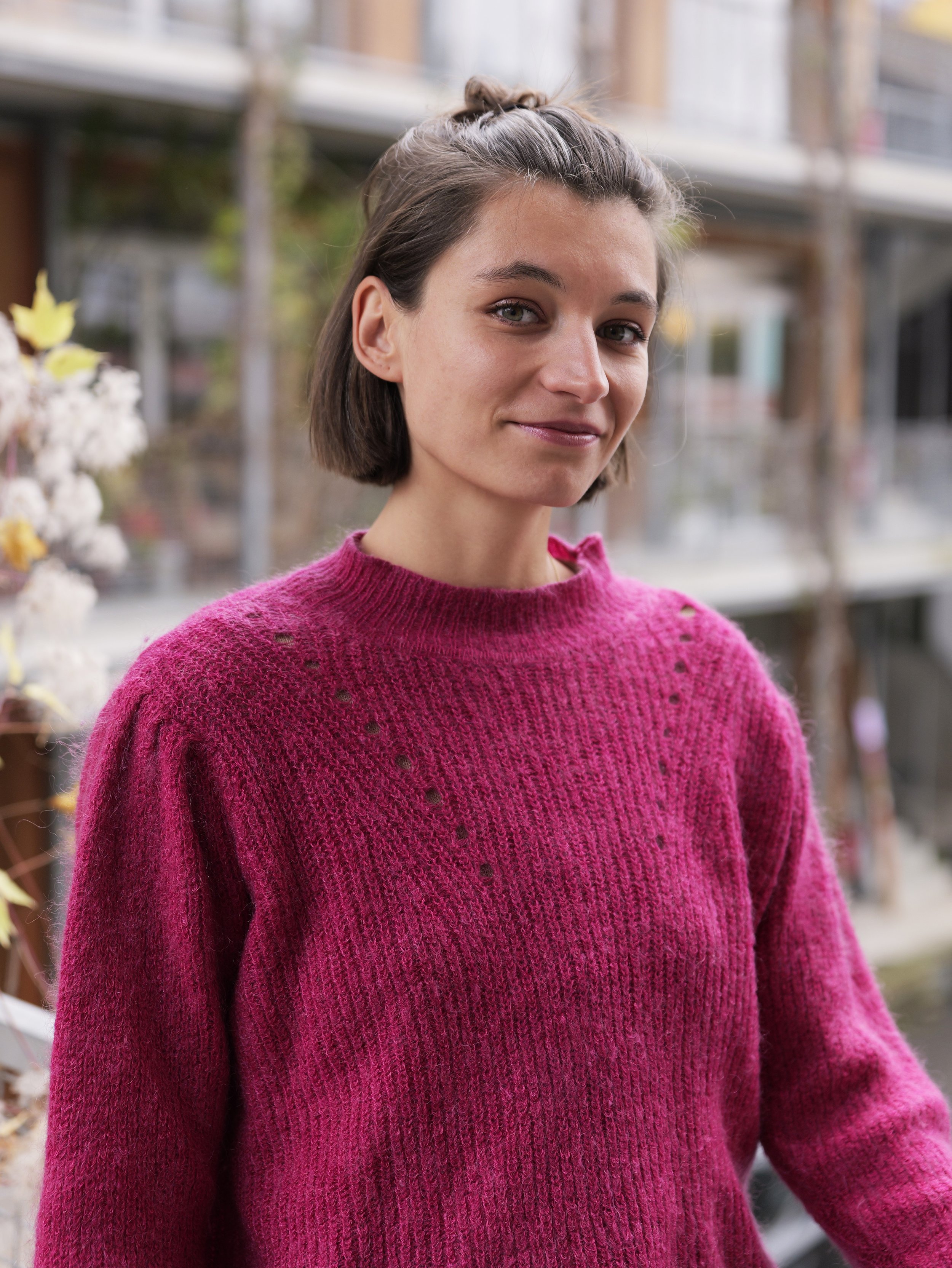 A woman with short dark hair wearing a pink sweater with small holes and long earrings, standing against a dark green background.