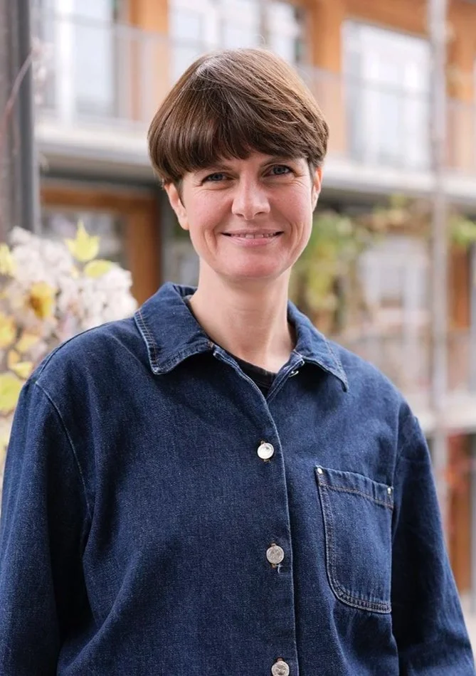 A woman with short brown hair, wearing a blue denim jacket, smiling outdoors with a building and plants in the background.