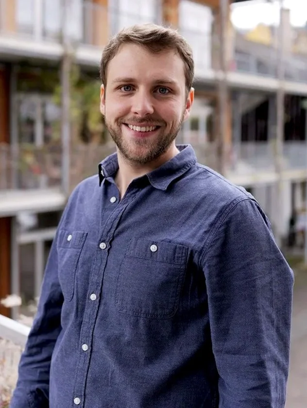 A smiling man with brown hair and beard standing outdoors in front of a multi-level apartment or condo complex with balconies. He's wearing a dark blue button-up shirt.