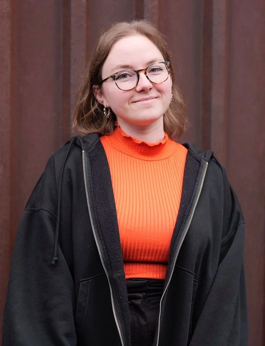A young woman with glasses and earrings, wearing an orange ribbed turtleneck top and a black hoodie, standing in front of a wooden background, smiling softly.