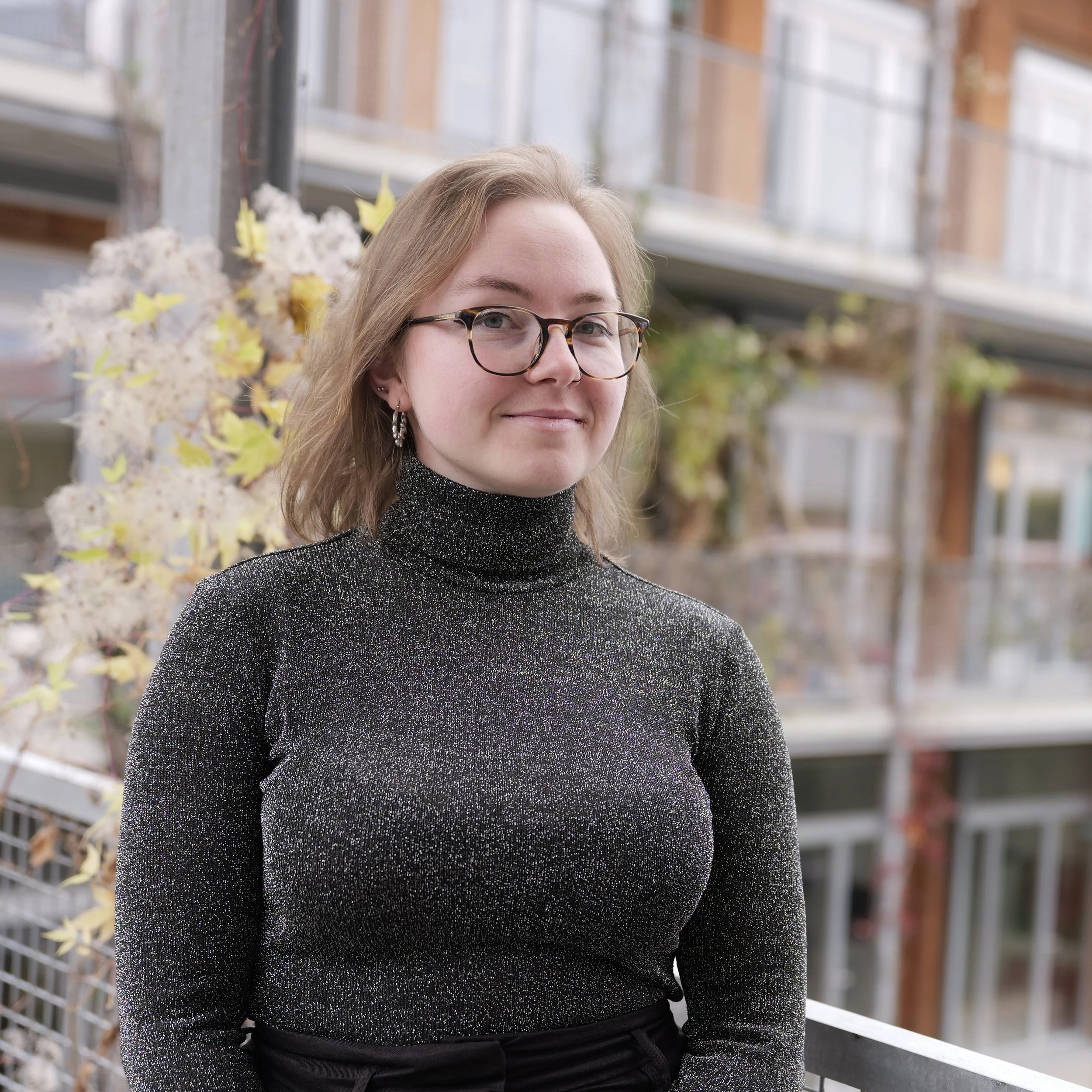 A young woman with glasses and earrings, wearing an orange ribbed turtleneck top and a black hoodie, standing in front of a wooden background, smiling softly.