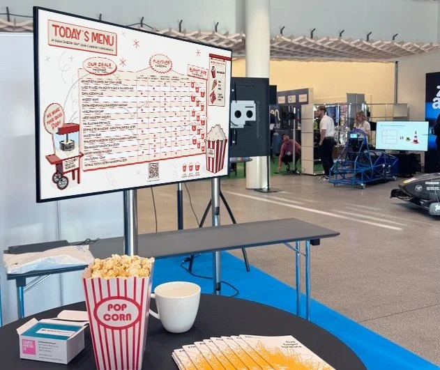 photograph of the Open-I fair. The stand of ETH student industry programmes. In the front som popcorn baskets and information handouts. In the back a screen showing the keyvisual