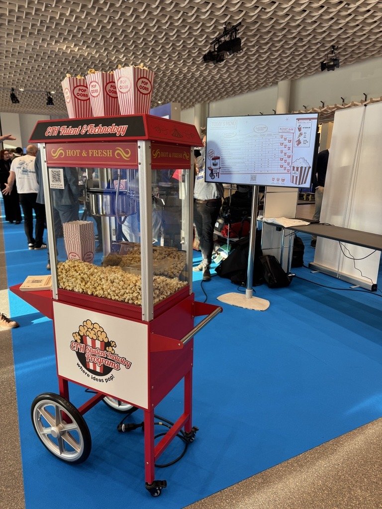 photograph of the Open-I fair. The stand of ETH student industry programmes. In the front a mobile popcorn machine. In the back a screen showing the Keyvisual