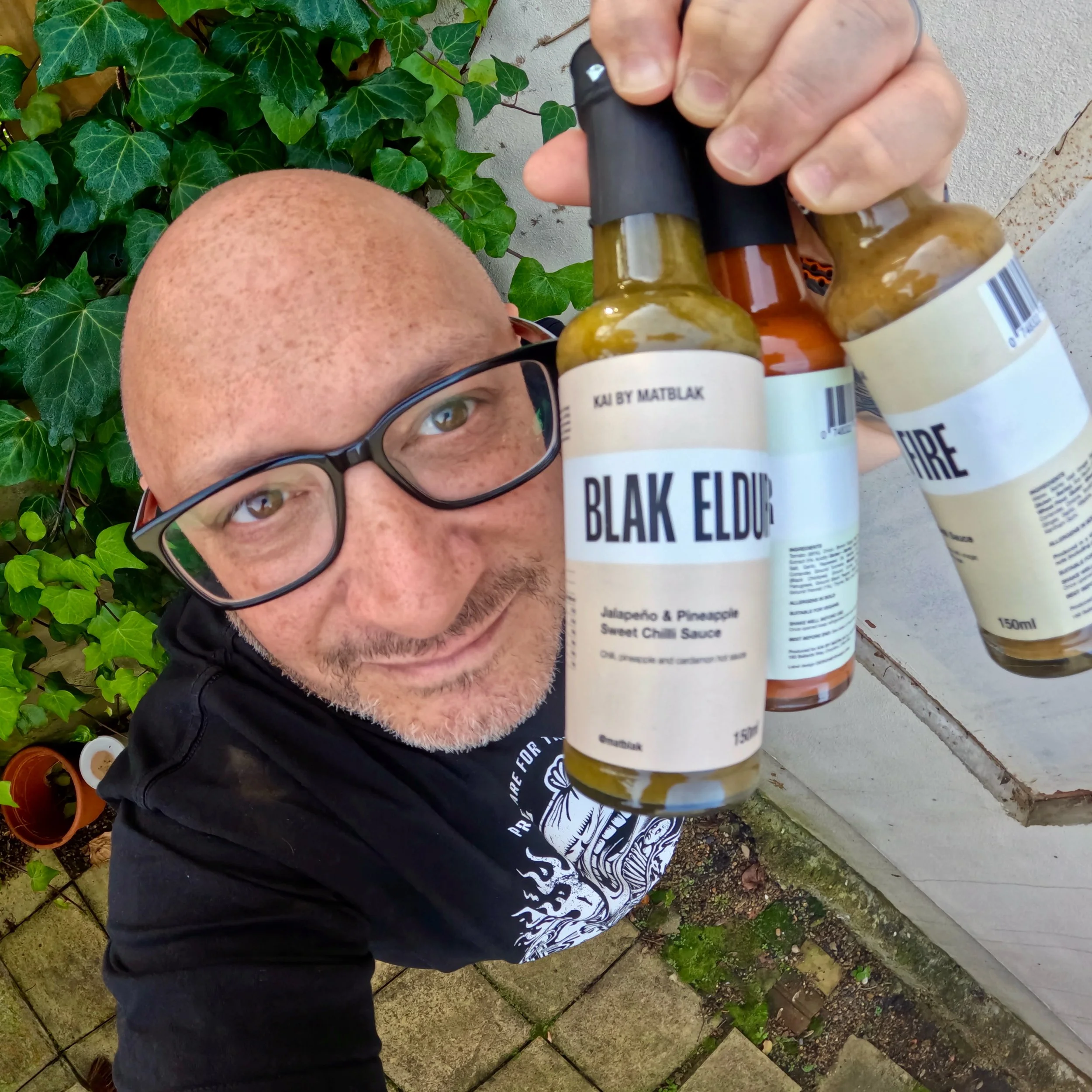 A man with glasses and a black t-shirt holding three bottles of chili sauce labeled "Blak Eldur" in front of him, standing outdoors near a wall with green ivy and potted plants.