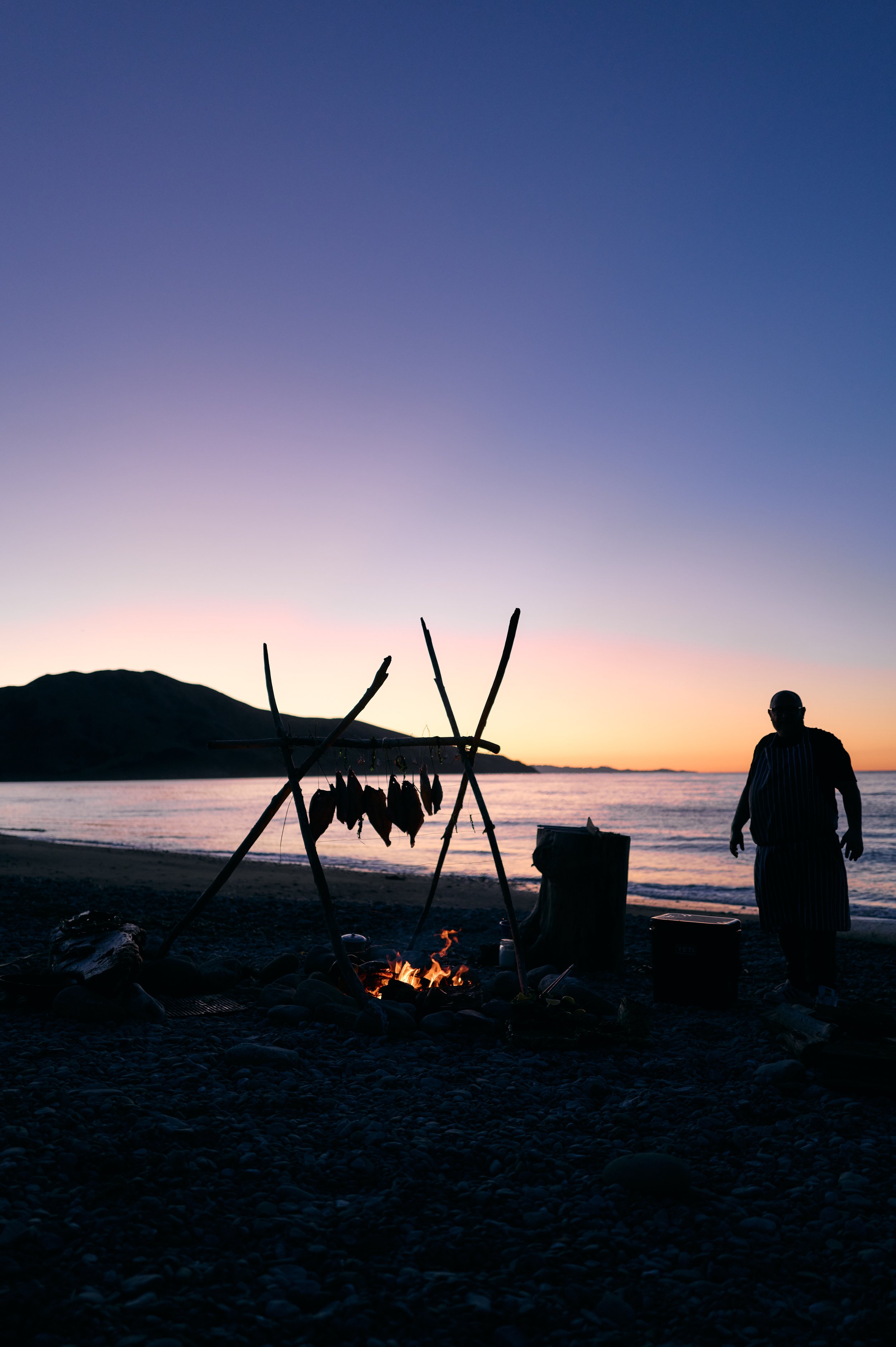 A person in a striped apron standing on a beach at sunset, near a campfire with hanging fish, with mountains and water in the background.