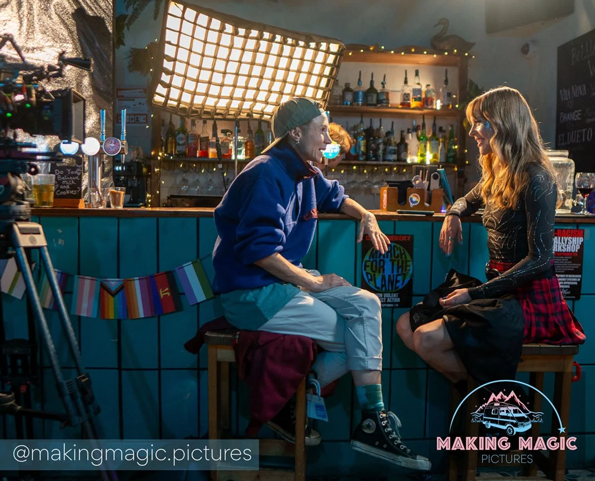 A bar with two people sitting and talking to each other, one is wearing a blue jacket and cap, the other a black top with patterns and a red plaid skirt, with bottles of alcohol on shelves in the background, and a large light fixture overhead.