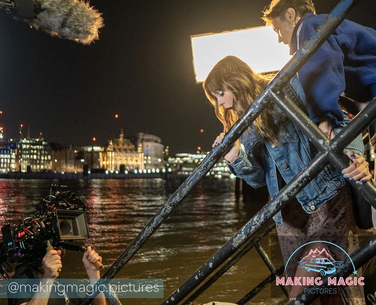 Magic and Jo are leaning over a metal railing and looking down. A film camera operator is capturing the scene.