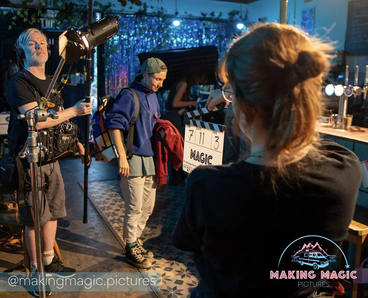 Filmmaker and crew filming a scene at a bar with colorful decor. A  person holds a clapperboard with the words "MAGIC" on it, while another person holds a light. 