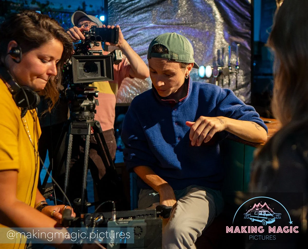 Film crew setting up shot in a green cap in a dimly lit venue with colorful lighting, behind a logo that says 'Making Magic Pictures'.