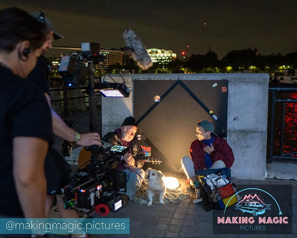Filming crew captures Magic and a busker playing guitar and singing at night, with a dog sitting between them, illuminated by a small light. A boom microphone hovers above.