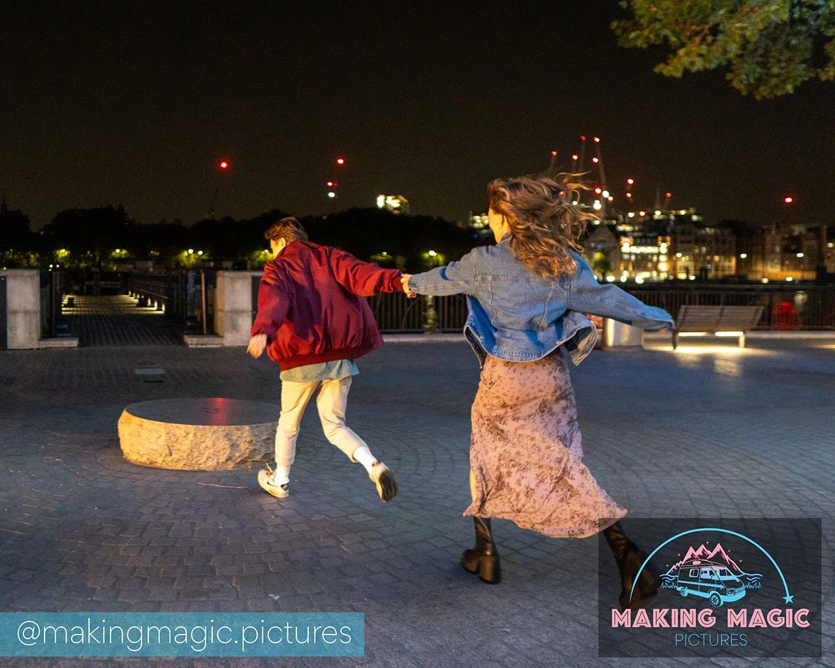 Magic and Jo hold hands & dance on a waterfront promenade at night. Jo has long, wavy hair and wears a denim jacket and floral skirt, while the Magic wears a red jacket and light-colored pants. City lights and buildings are visible in the background.