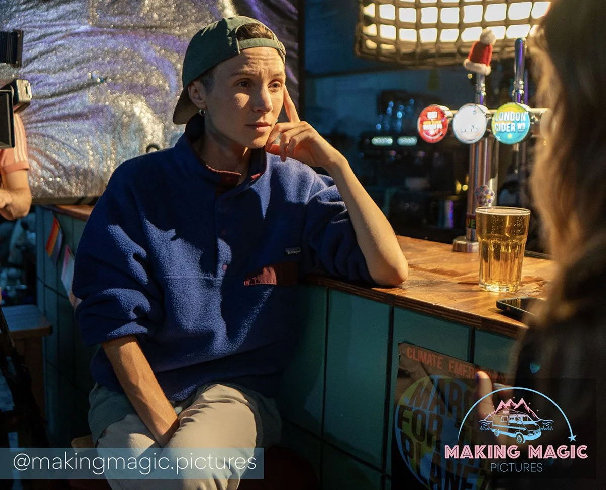Magic (Dom PC), wearing a backwards cap and a blue fleece jacket, sitting at a bar with a glass of beer, looking thoughtful. The bar has several beer taps and a neon sign in the background.